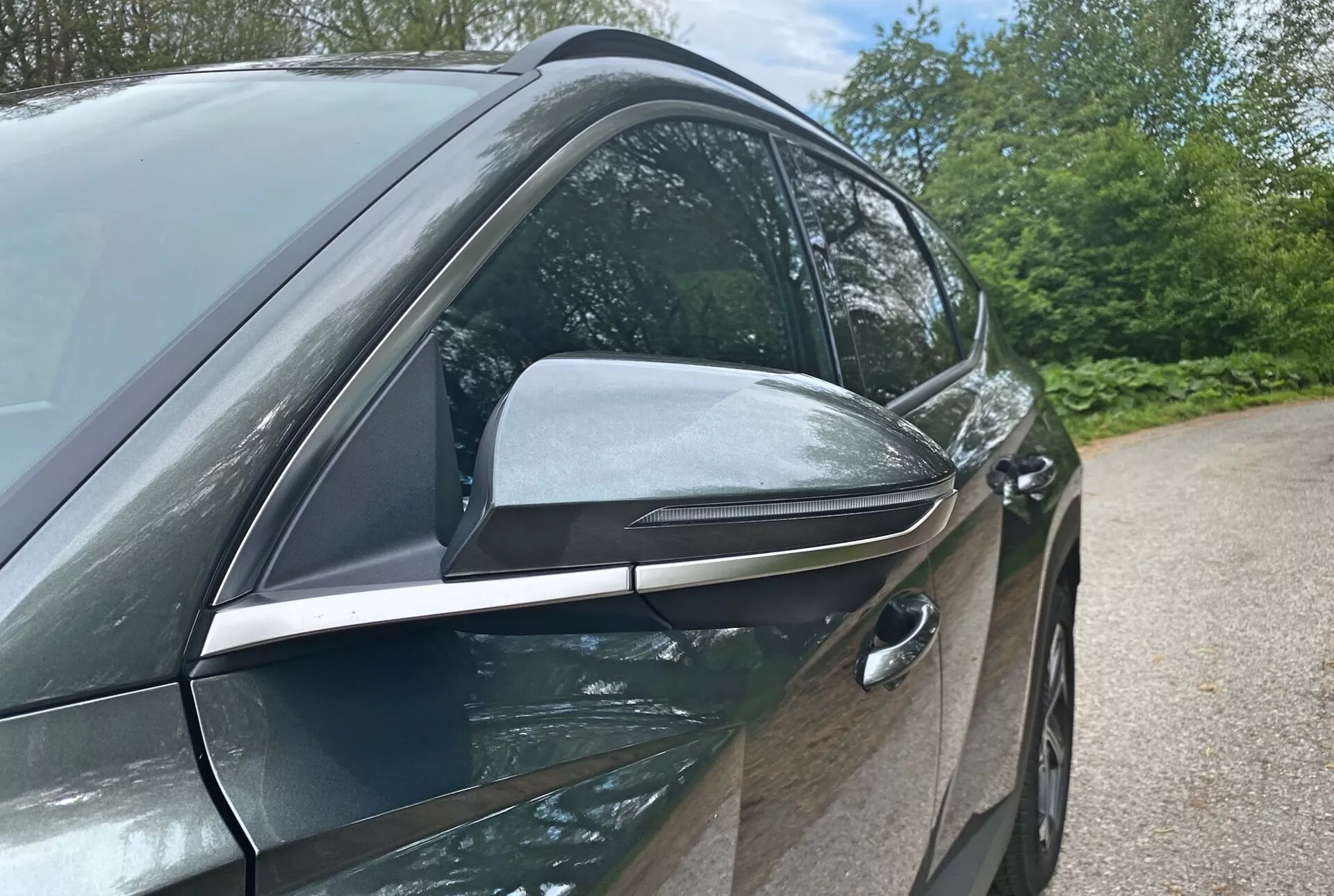 Zbliżony widok boczny Hyundai Tucson 2020 1.6 T-GDI Pure podkreślający jego nowoczesny design i zaawansowane funkcje technologiczne z naciskiem na stronę kierowcy. Zdjęcie wykonane z niskiego kąta uwydatnia eleganckie, aerodynamiczne lusterko boczne z zintegrowanymi kierunkowskazami i czarnymi akcentami, sugerując zaawansowane funkcje. Wbudowana klamka przednich drzwi od strony kierowcy wskazuje na system bezkluczykowego dostępu, przyczyniając się do aerodynamicznej wydajności pojazdu. Błyszczące, w kolorze nadwozia drzwi oraz chromowane wykończenia okien sugerują pakiet premium. Elementy tła, takie jak drzewa i niebo, sugerują ustawienie na zewnątrz.