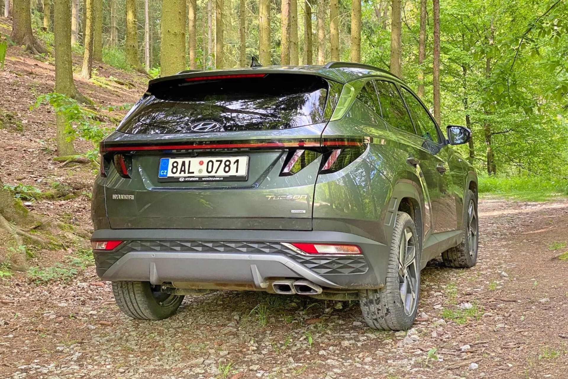 Zdjęcie nowoczesnego SUV-a Hyundai Tucson 2020 w leśnym otoczeniu na żwirowej drodze, uchwycone z tylnego, trzy czwarte kąta. Zdjęcie podkreśla prawą stronę i tylny profil pojazdu z widocznymi tylnymi drzwiami, tylnymi światłami LED, podwójnym układem wydechowym, tylnym spojlerem i felgami aluminiowymi.
