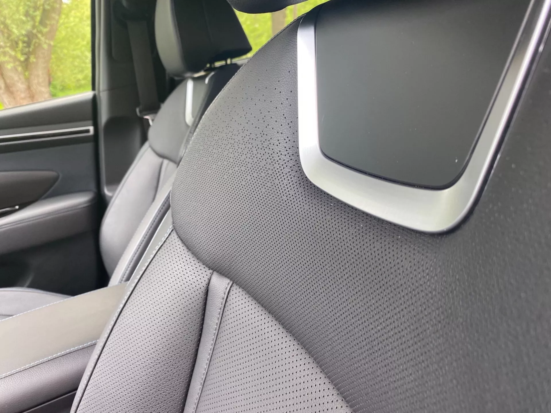 Zbliżenie wnętrza Hyundai Tucson 2020, skoncentrowane na prawym słupku A i części przedniego prawego siedzenia. Słupek A jest pokryty teksturowanym materiałem, prawdopodobnie tkaniną lub skórą syntetyczną, sugerując nacisk na estetykę lub redukcję hałasu. Siedzenie ma dwukolorowy design i jest perforowane dla wentylacji. Zauważalna cecha przy skrzyżowaniu słupka A i podsufitki może być częścią systemu audio, komponentem poduszki powietrznej lub czujnikiem wspomagania kierowcy, podkreślając zaawansowane funkcje bezpieczeństwa lub technologii.