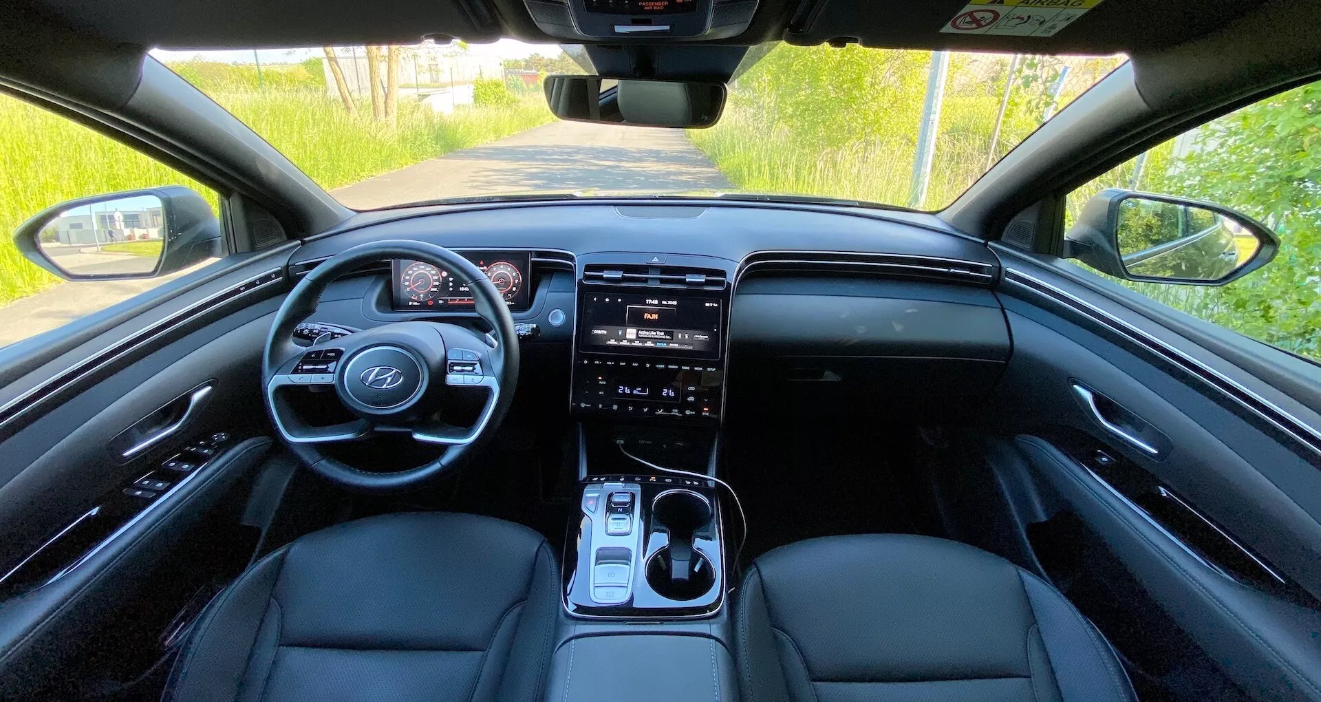 Widok wnętrza Hyundai Tucson 2020 z tylnego siedzenia ukazujący przednią deskę rozdzielczą, kierownicę z wbudowanymi przyciskami, cyfrowy panel wskaźników, duży system informacyjno-rozrywkowy z ekranem dotykowym, przyciski sterowania klimatyzacją, dźwignię zmiany biegów oraz przednie siedzenia wyściełane skórą w naturalnym świetle dziennym.