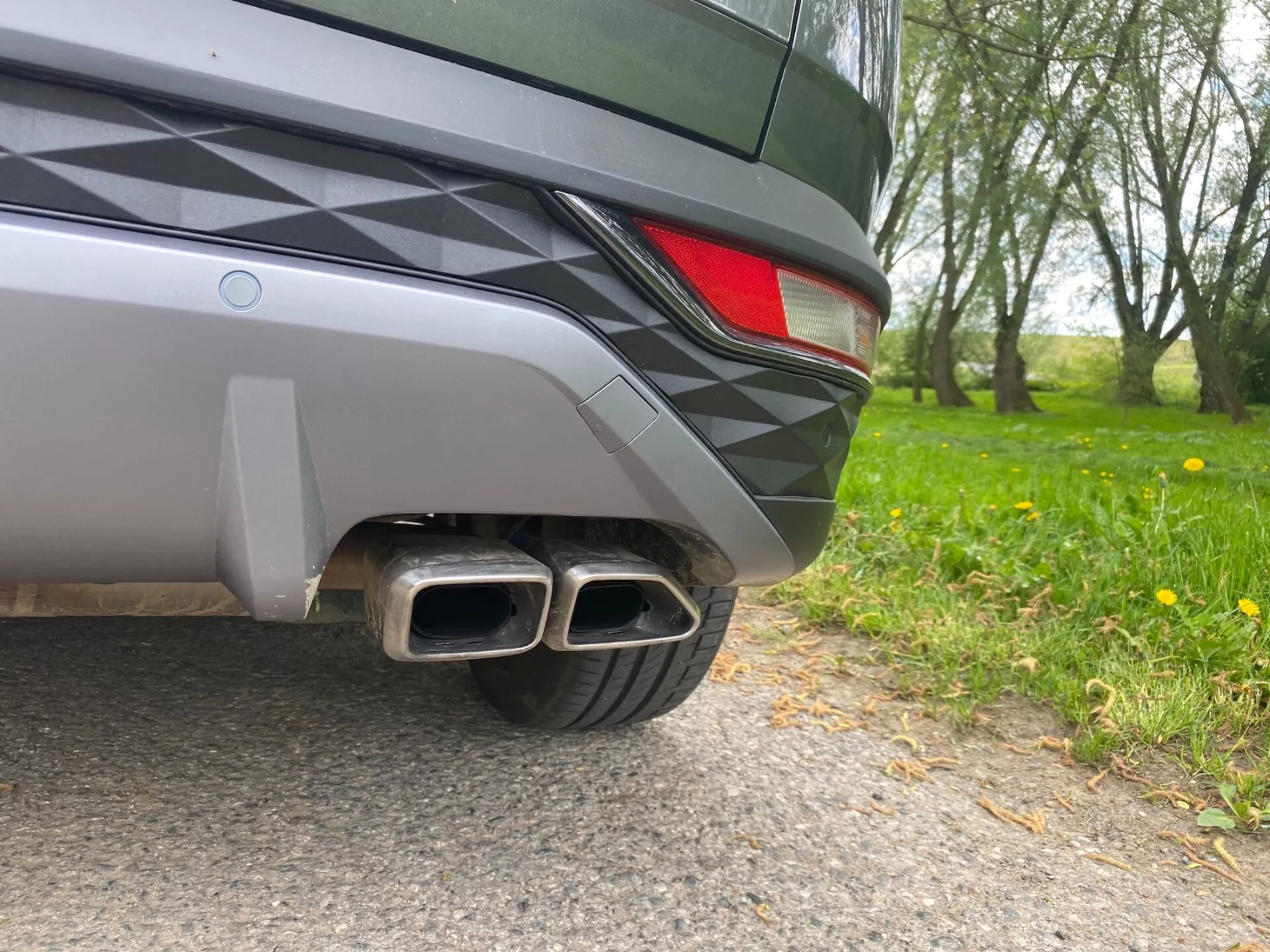 Zbliżenie tylnej części Hyundaia Tucson 2020, ujęcie wykonane z poziomu ziemi. Widoczne szczegóły obejmują dwukolorowy tylny zderzak, fragment tylnej lampy LED oraz podwójne metalowe rury wydechowe z prostokątnymi końcówkami, co sugeruje sportowy wygląd. Elegancki design pojazdu i metaliczna farba odbijają otaczającą przyrodę.