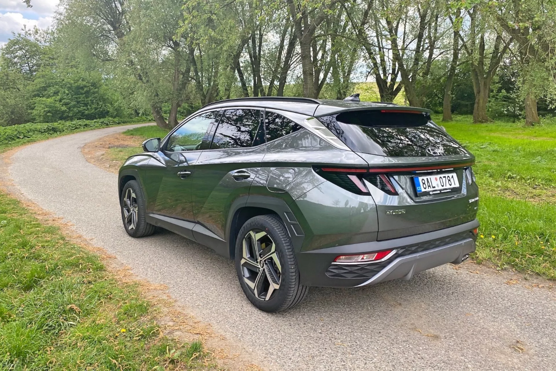Obraz przedstawia nowoczesny crossover SUV z tylnego bocznego kąta, podkreślając elegancki, aerodynamiczny wygląd i nachyloną linię dachu. Ciemnozielony pojazd posiada tylną klapę w stylu liftback, sportowy spojler i duże felgi aluminiowe z wieloszprychowym wzorem. Widoczne elementy to nowoczesne tylne światła LED, podwójne rury wydechowe i tylna wycieraczka. SUV jest zaparkowany na żwirowej ścieżce otoczonej zielenią, co sugeruje park lub wiejskie otoczenie.