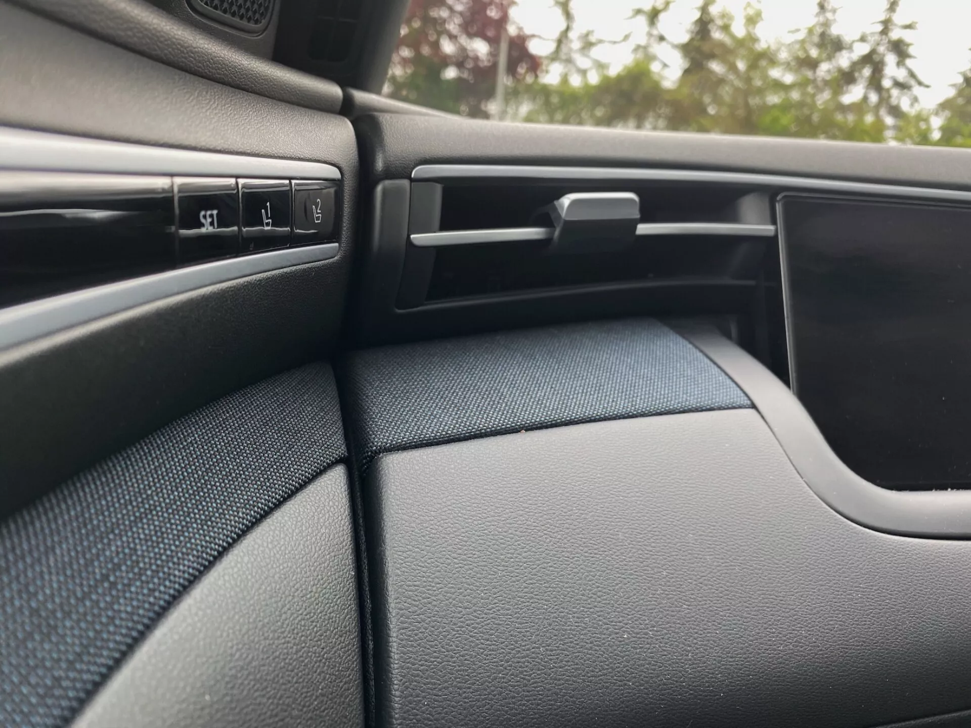 Szczegół wnętrza Hyundai Tucson 2020, ukazujący ciemny, teksturowany bok deski rozdzielczej po stronie pasażera i część panelu drzwi. Scena zwraca uwagę na kilka przycisków kontrolnych, regulowany nawiew z poziomymi żaluzjami, zamykaną schowek i zakrzywiony design panelu drzwi, który płynnie łączy się z deską rozdzielczą.