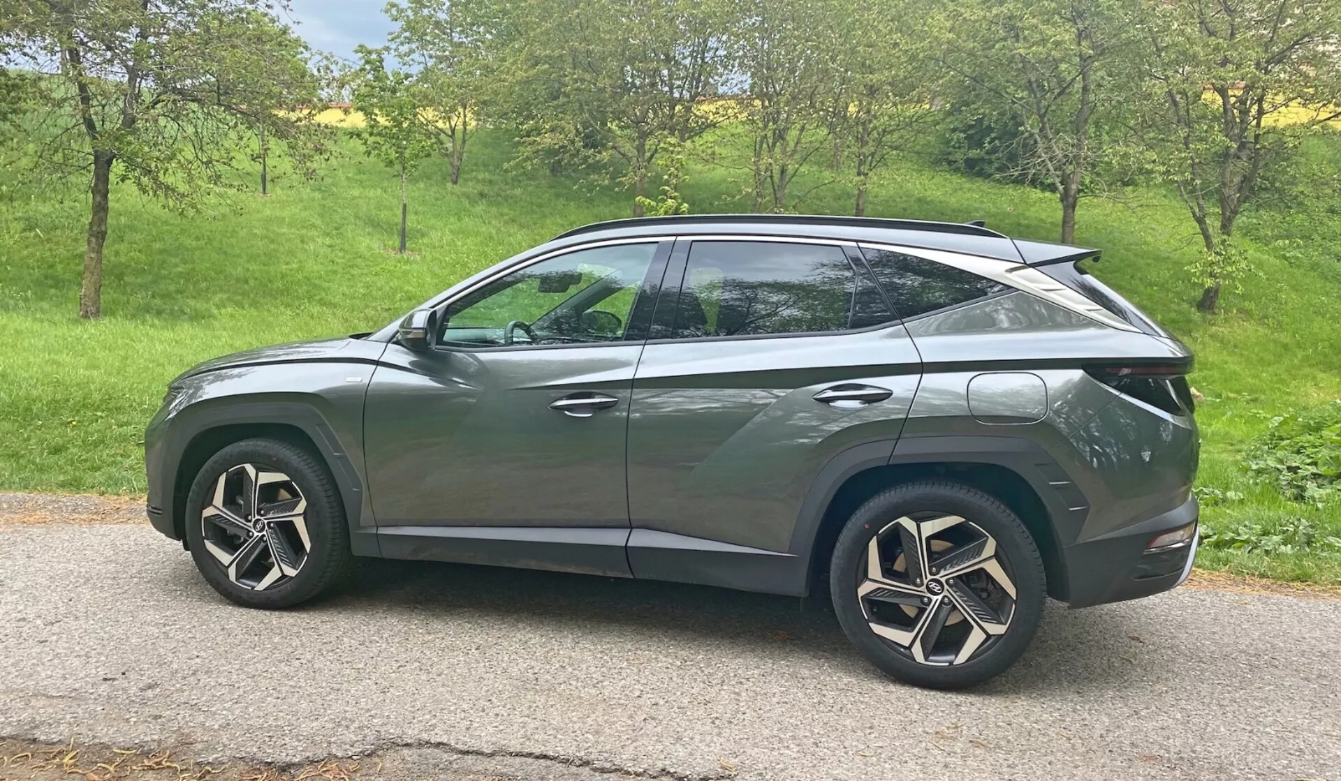 Boczny profil SUV-a Hyundai Tucson 1.6 T-GDI Pure 2020 w naturalnym świetle dziennym. Pojazd znajduje się na trawiastym terenie z drzewami w tle, co sugeruje wiejskie lub podmiejskie otoczenie. Nowoczesne cechy projektowe obejmują reflektory LED, zintegrowane kierunkowskazy na lusterkach bocznych, przyciemniane szyby, aerodynamiczne aluminiowe felgi i pochylony dach w stylu coupé. Elegancka karoseria z charakterystycznymi liniami i ukrytymi klamkami drzwi sugerują zaawansowany system bezkluczykowy, podczas gdy tylny spojler wskazuje na aerodynamiczne udoskonalenia. Samochód jest centralnie umieszczony w ramce, zapewniając przejrzysty, niezakłócony widok z perspektywy średniej wysokości.
