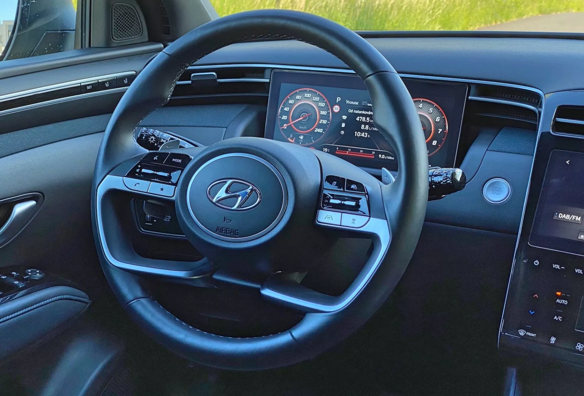 Wnętrze Hyundai Tucson 1.6 T-GDI 2020 z perspektywy kierowcy, skupiające się na nowoczesnej, trójramiennej kierownicy z logo Hyundai, przyciskach sterowania mediami i tempomatem, częściowo widocznym zestawie wskaźników z prędkościomierzem i obrotomierzem oraz centralnym systemie infotainment na desce rozdzielczej.