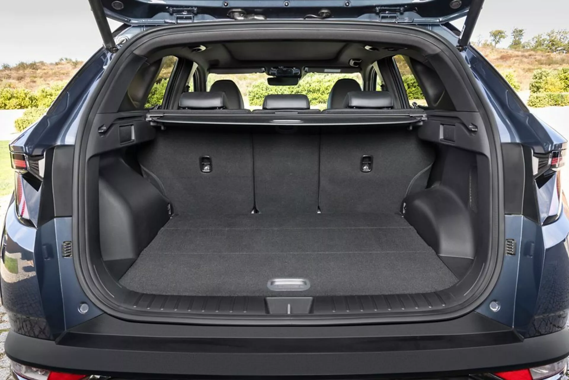 Tylna przestrzeń bagażowa Hyundai Tucson 2020 z otwartą klapą bagażnika, prezentująca wykładzinę na płaskiej podłodze ładunkowej, punkty mocowania, schowki, gniazdo zasilania 12V, składane siedzenia tylne, chowany pokrowiec bagażowy, trwałe plastikowe ściany boczne oraz widok na tylny zderzak, na zewnątrz.