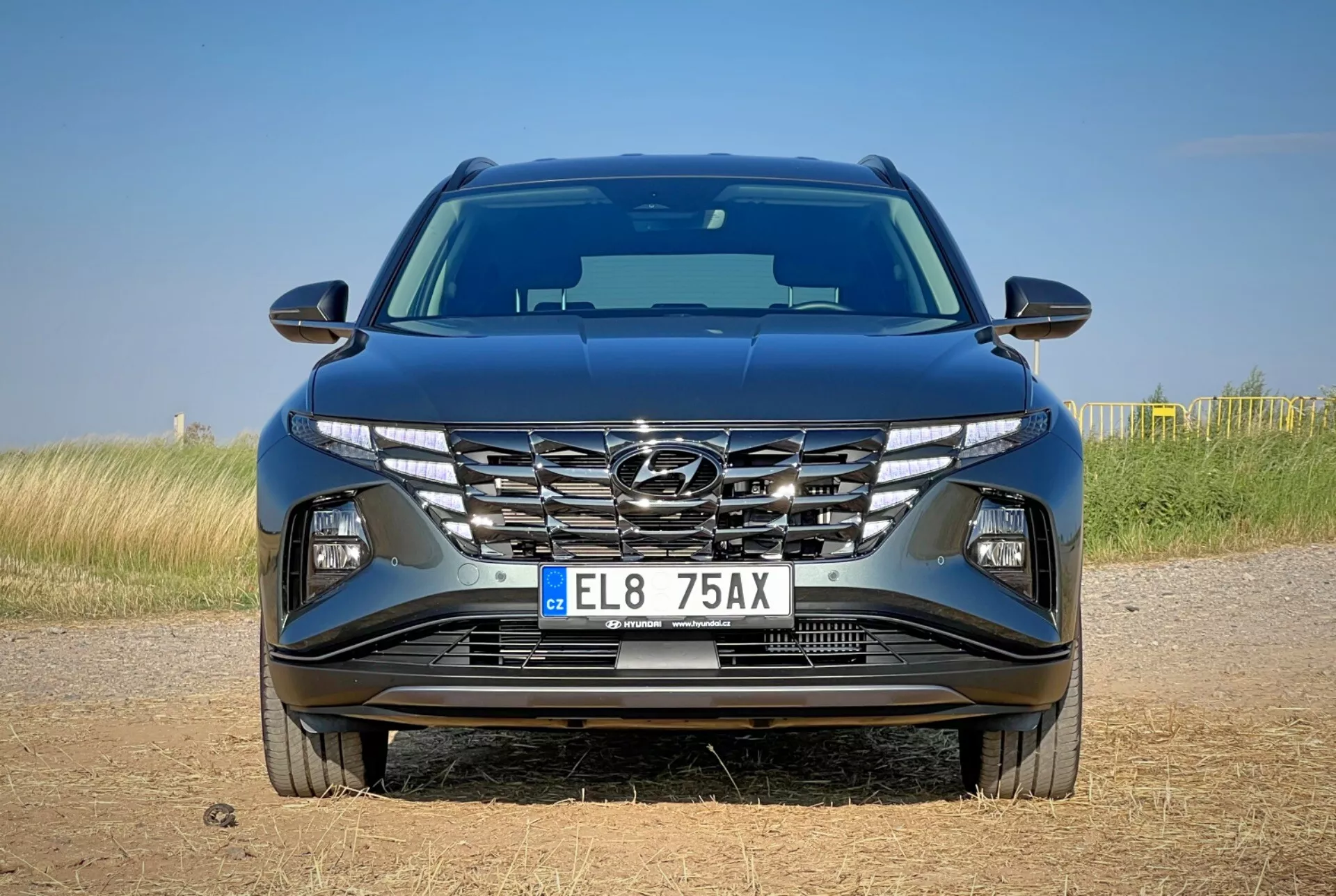 Przedni widok Hyundai Tucson 2020 z aerodynamicznym designem maski, kaskadowym grillem, ostrymi reflektorami LED i szczegółowym zderzakiem z możliwymi funkcjami technologicznymi, ustawiony na tle naturalnego krajobrazu