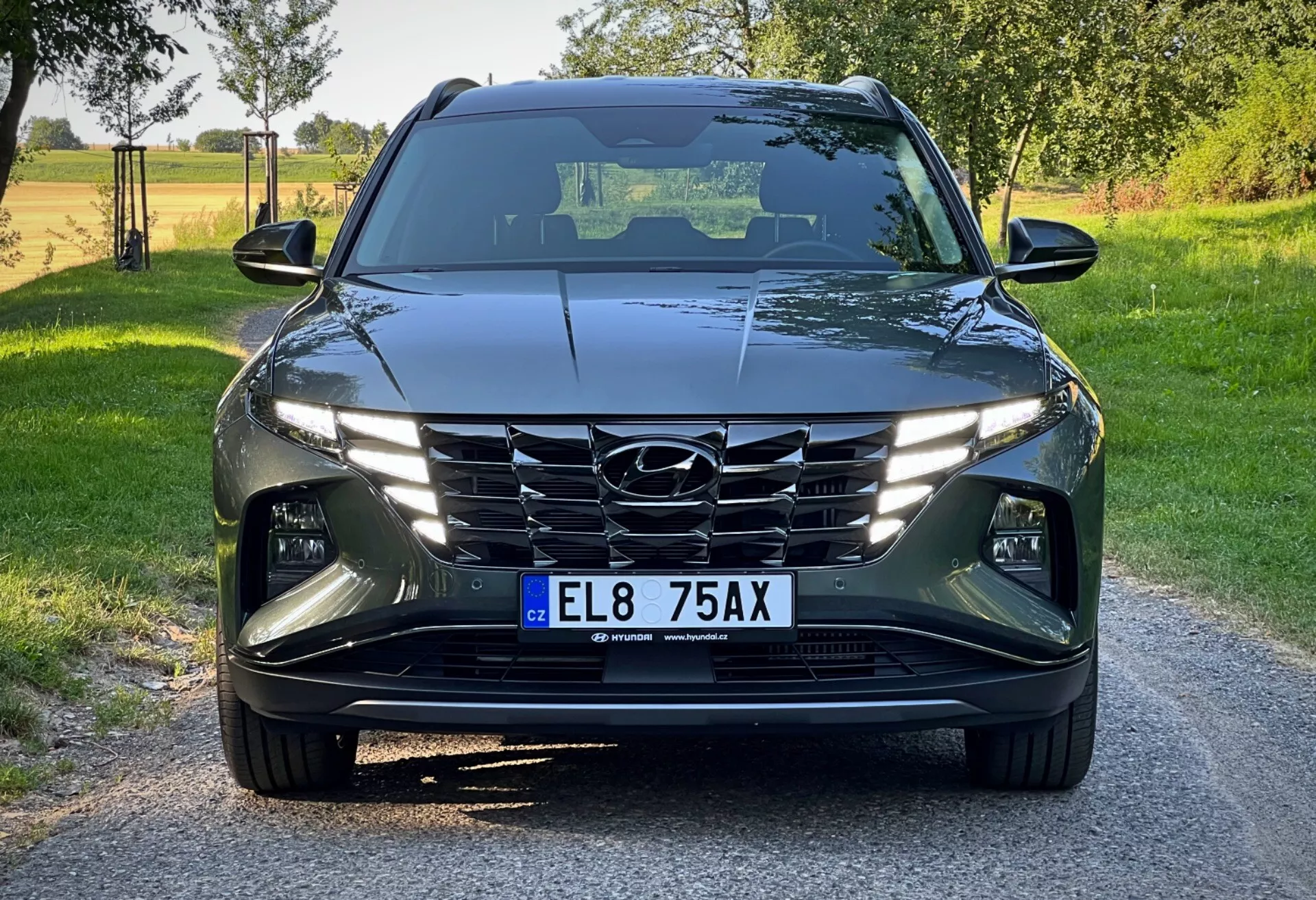 Hyundai Tucson 2020 widziany z niskiej perspektywy frontowej, ukazujący jego nowoczesny design z ostrymi, kątowymi liniami, reflektorami LED, kaskadową atrapą chłodnicy i zaawansowanymi systemami wspomagania kierowcy na tle jasnego, wiejskiego dnia