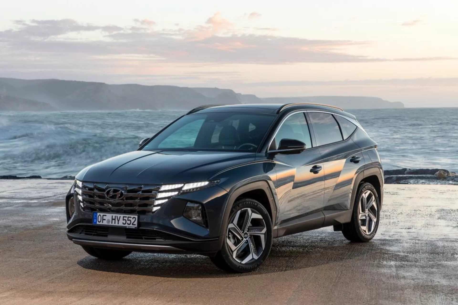 Nowoczesne SUV Hyundai Tucson 2020 zaparkowane na plaży z oceanem i horyzontem w tle, prezentujące widok z przodu i boku pojazdu, co podkreśla jego design, charakterystyczną kratkę z logiem, kątowe reflektory LED i sportowe felgi aluminiowe