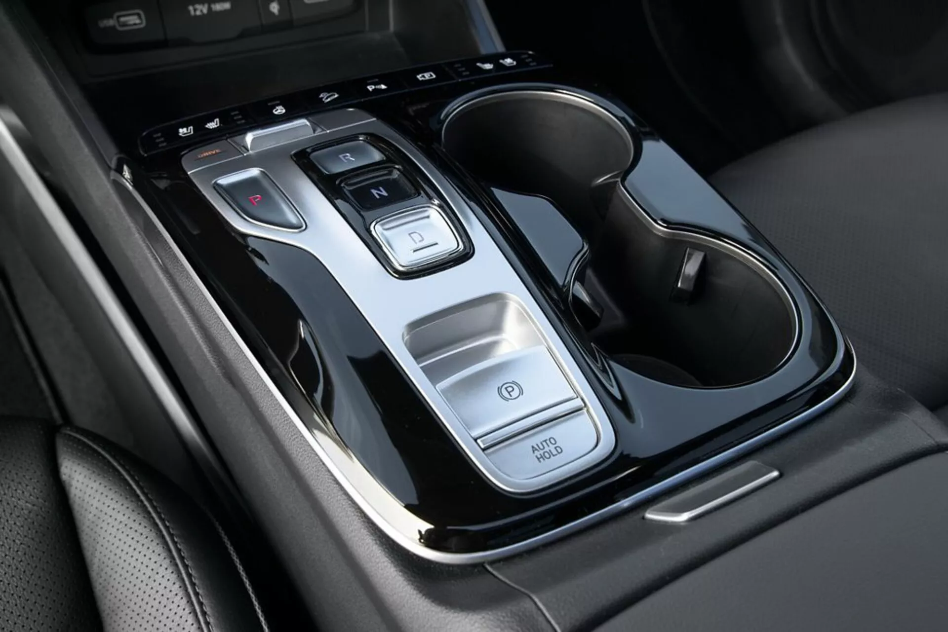 Zbliżenie na konsolę środkową Hyundai Tucson 2020 z dźwignią zmiany biegów automatycznych, przyciskami sterowania, funkcją Auto Start/Stop, przełącznikiem świateł awaryjnych i podwójnym uchwytem na napoje z akcentami chromowanymi, z górnego widoku z lekkim nachyleniem.