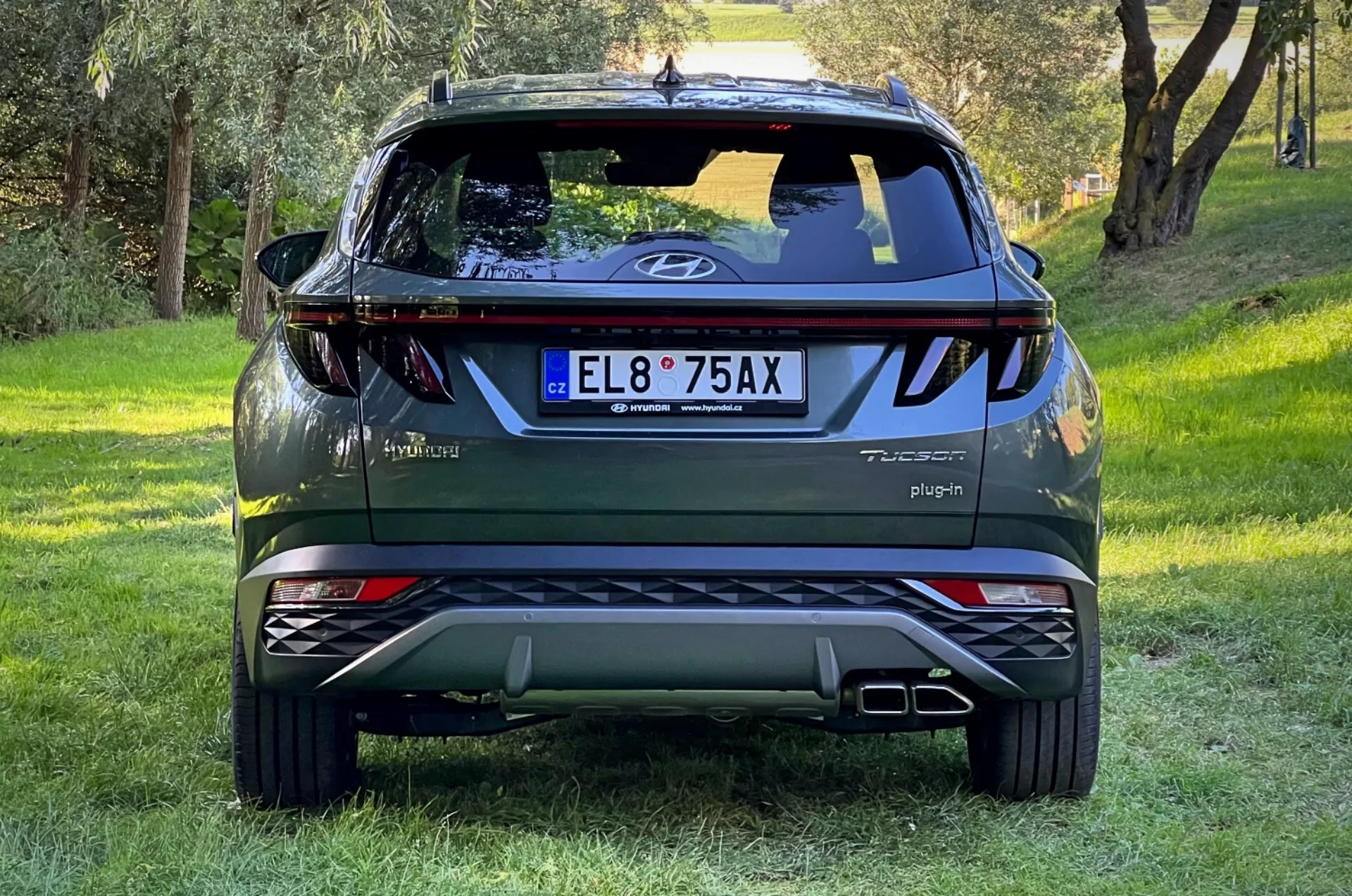 Tylny widok SUV-a Hyundai Tucson 2020 na trawiastym stoku z leśnym tłem, pokazujący logo na klapie bagażnika, odznaki Tucson i Hybrid, napis Plug-in, odblaskowe światła, dynamiczne tylne światła, tylną wycieraczkę, wysoko umieszczony światło stop, podwójne końcówki wydechu, srebrne płyty ochronne i odznakę AWD.