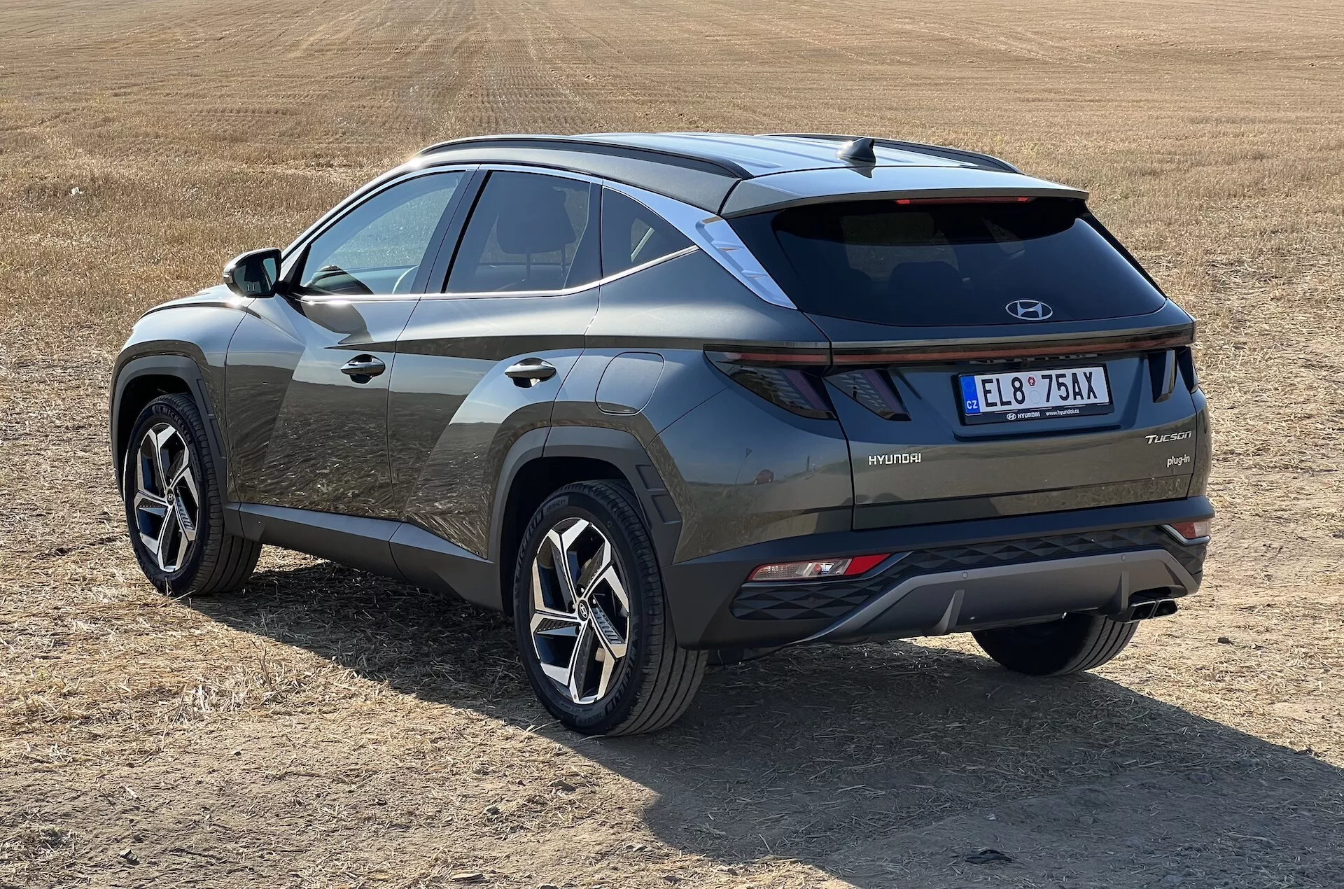 Nowoczesny SUV Hyundai Tucson 2020 w ukośnym widoku prezentujący profile tylne i boczne z oznaczeniem technologii hybrydowej na suchym trawiastym polu, podkreślający tylne światła LED, wstawkę zderzaka w kontrastowym kolorze i sportowe koła o wielu szprychach