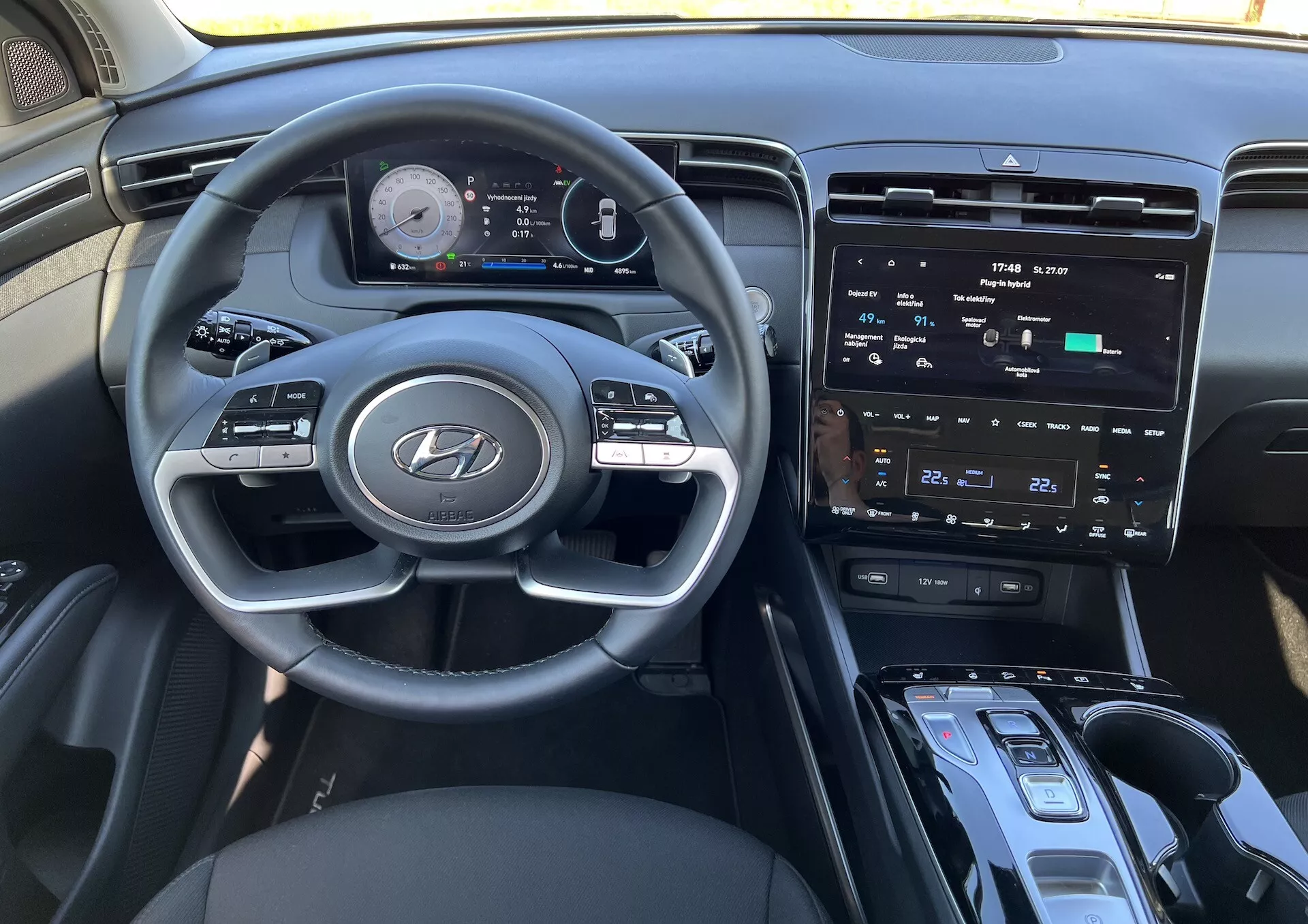 Wnętrze Hyundai Tucson 2020 przedstawiające kierownicę z logo Hyundai, zaawansowany cyfrowy zestaw wskaźników, centralny dotykowy ekran systemu rozrywki, sterowanie klimatyzacją, przycisk start/stop, dźwignię zmiany biegów i możliwą strefę ładowania bezprzewodowego z perspektywy kierowcy