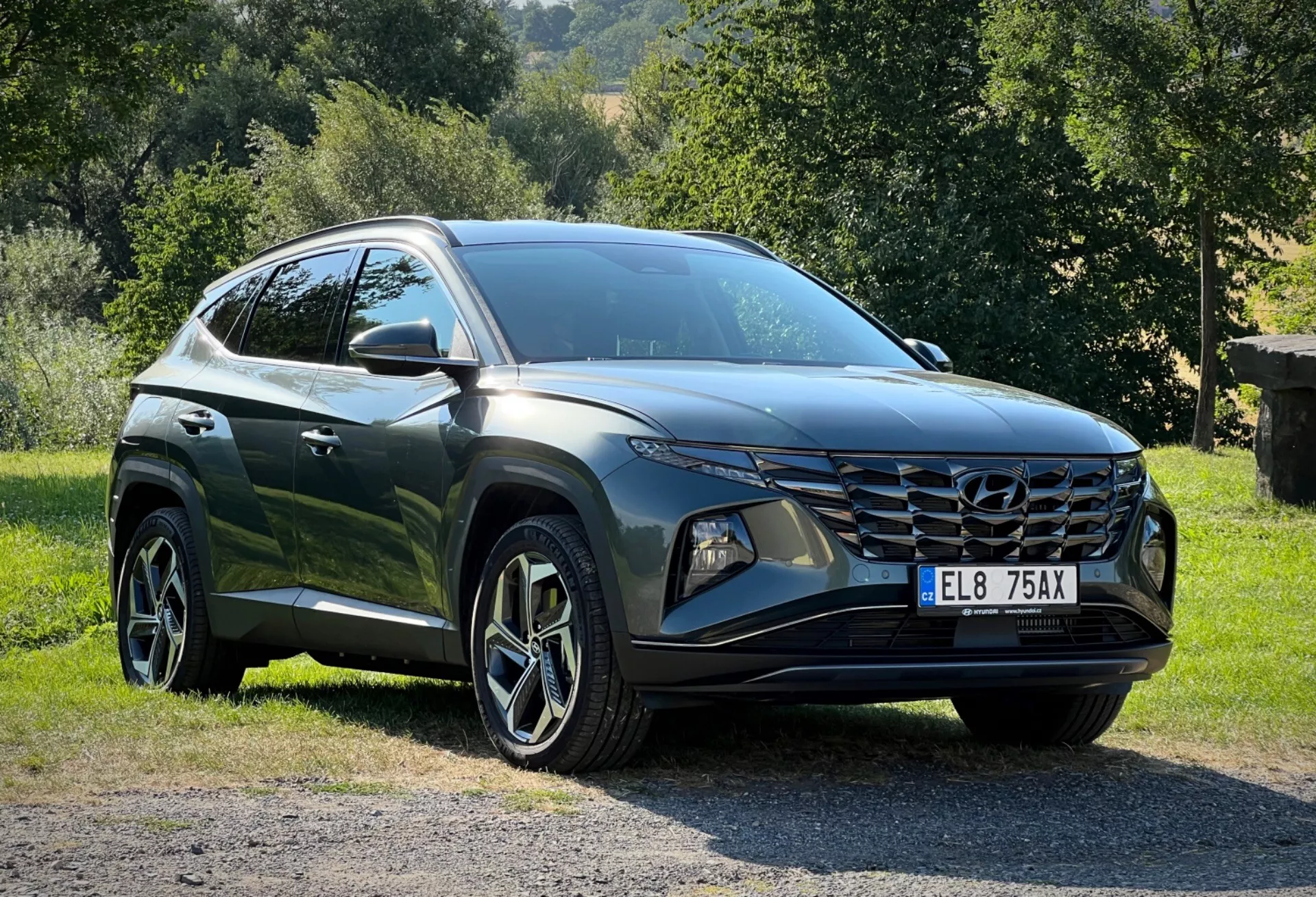 Hyundai Tucson 2020 zaparkowany na trawie z wiejskim tłem, prezentujący perspektywę czołową z trzech czwartych dla szczegółowego widoku jego eleganckich reflektorów LED, kątowej przedniej kratki i błyszczącego ciemnego wykończenia