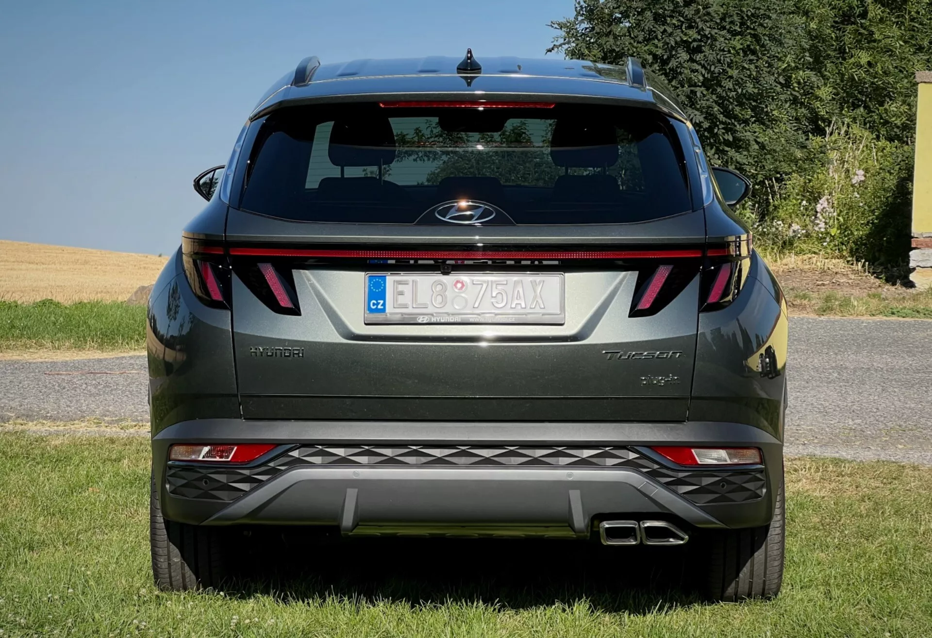 Tylny widok Hyundai Tucson 2020 pokazujący poziome tylne światła, centralną odznakę, nazwę wersji wyposażenia, zintegrowane czujniki parkowania na zderzaku i podwójny układ wydechowy, zaparkowany na obszarze otoczonym trawą przeciwko jasnemu niebu