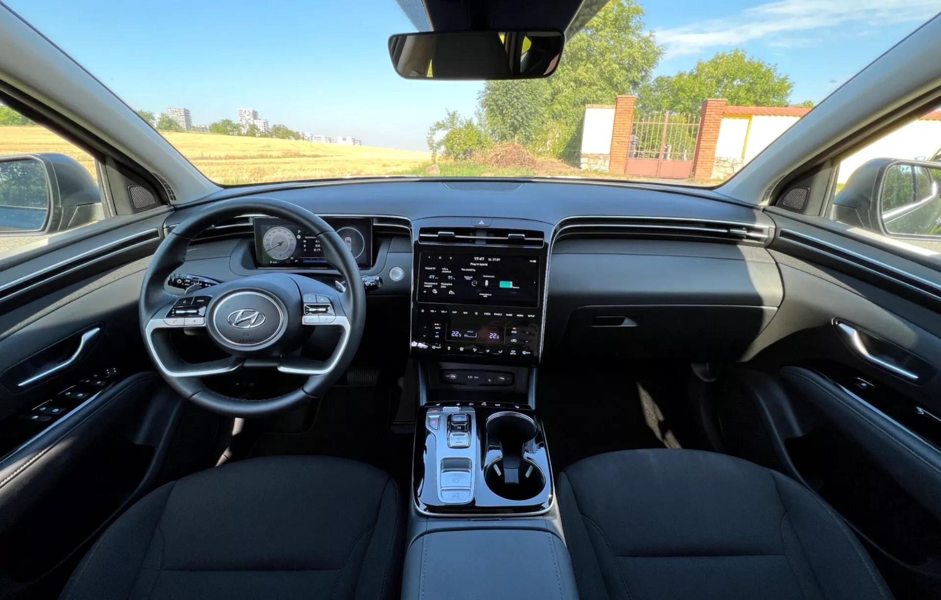 Wnętrze Hyundai Tucson 2020 przedstawiające przednie fotele, kierownicę z logo producenta i przyciskami sterowania, cyfrowy zestaw wskaźników, pionowy system infotainment na konsoli środkowej, automatyczną dźwignię zmiany biegów oraz dwutonowe wzornictwo deski rozdzielczej w widoku na poziomie oczu przypominającym perspektywę kierowcy