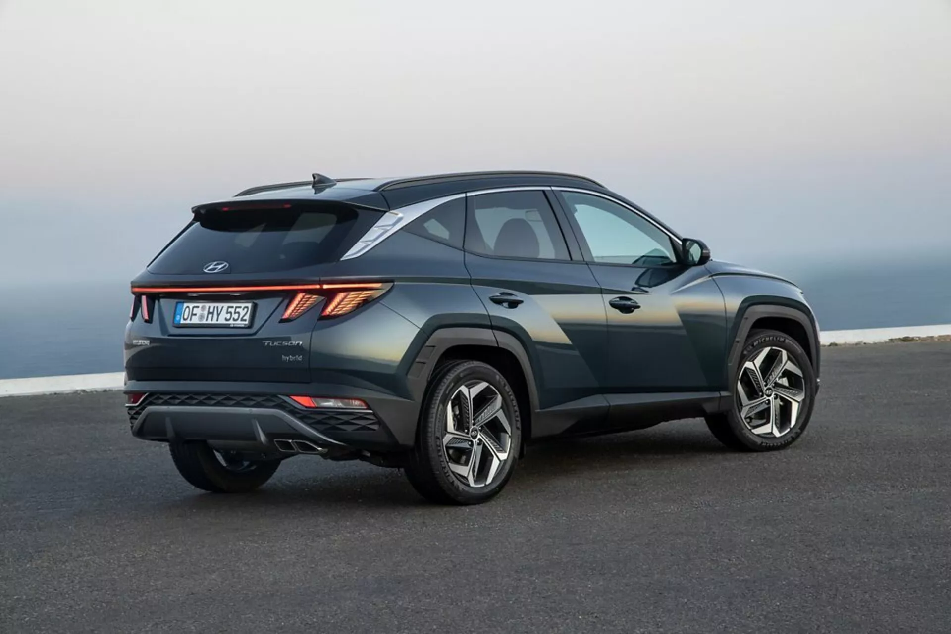 SUV Hyundai Tucson 2020 zaparkowany na gładkim asfalcie z krajobrazem nadbrzeżnym lub wzniesionym w tle, prezentujący jego elegancki, sportowy design, wzrastającą linię pasa, opadającą linię dachu, ostre zagięcia nadwozia, kątowe tylne światła LED, tylny spojler, zintegrowany dyfuzor, podwójne końcówki wydechu i duże felgi aluminiowe
