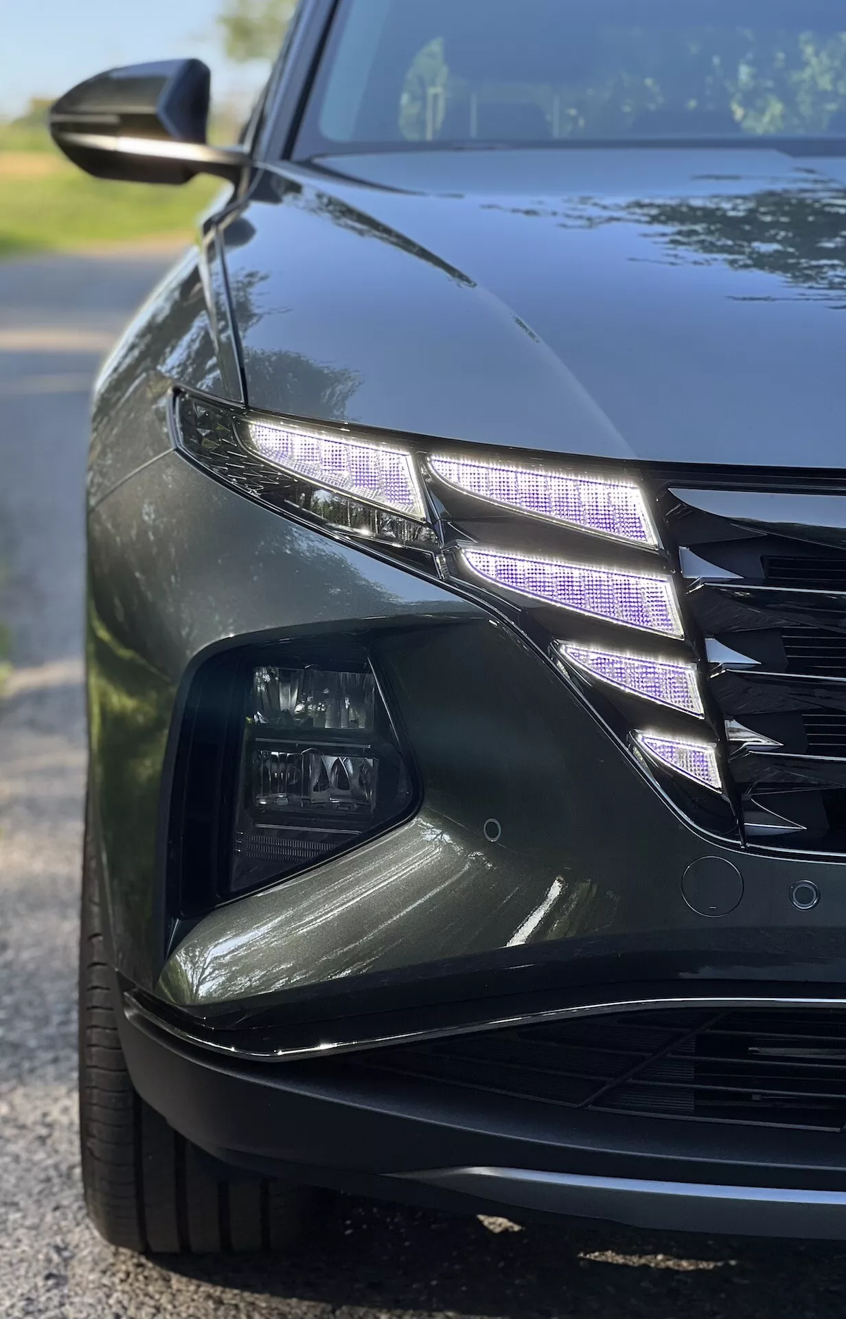 Zbliżony, kątowy widok na Hyundai Tucson 2020 przedstawiający skomplikowany projekt świateł LED i część kraty z połyskującym wykończeniem w warunkach słabego oświetlenia, ilustrujący cechy luksusowych samochodów i nowoczesną technologię oświetleniową samochodów.