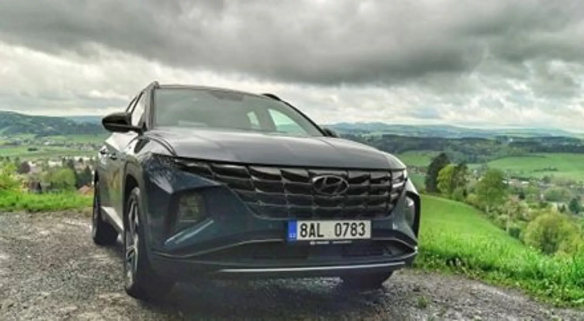 SUV Hyundai Tucson 2020 zaparkowany na drodze żwirowej, prezentujący śmiałą kratę, eleganckie reflektory LED i metaliczny lakier w dynamicznym widoku z niskiego kąta przedniego, z bujnym, pagórkowatym krajobrazem i pochmurnym niebem w tle
