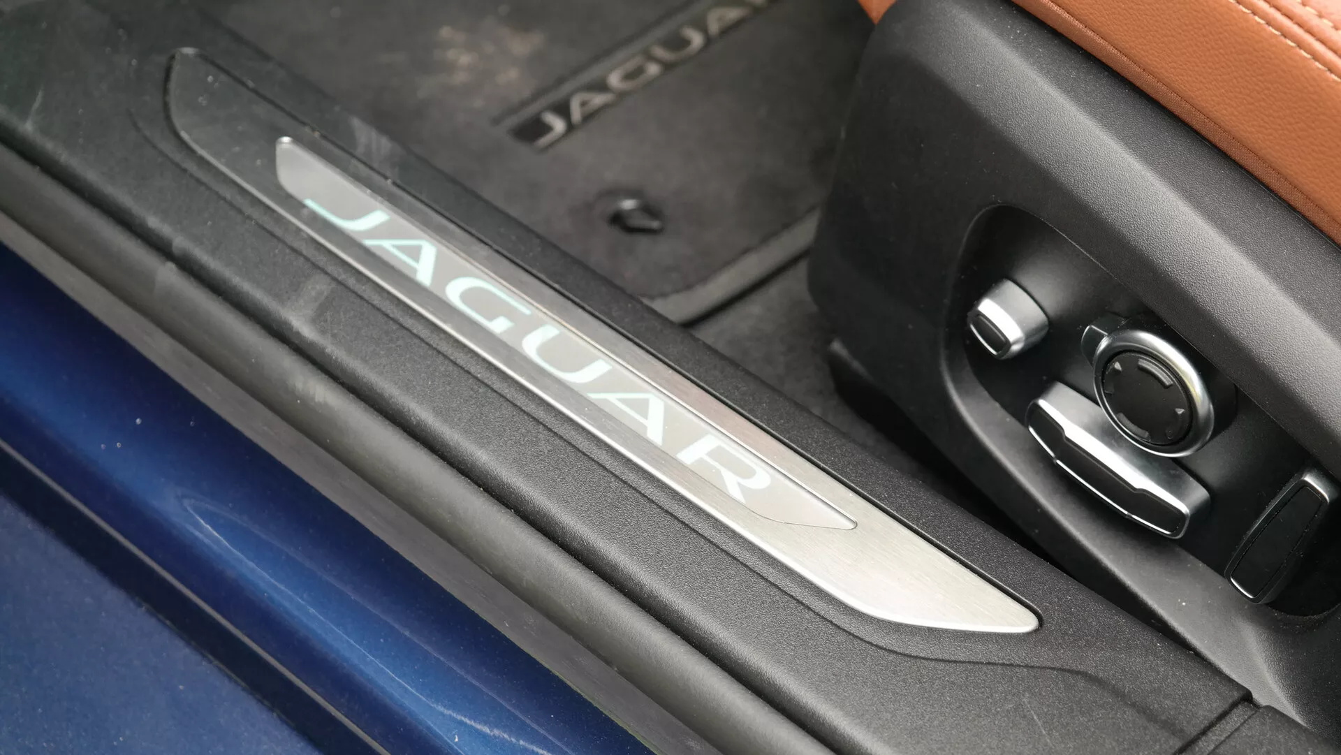 Zbliżenie szczegółu wnętrza Jaguara XF 2021, podkreślające podświetlany znak 'JAGUAR' na szczotkowanej metalowej listwie progowej, obok wzoru okręgów i przycisku kontrolnego. Kontrastujące tekstury obejmują chromowe wykończenie, zarys niebieskiego nadwozia i górną część z skóry w luksusowym, nowoczesnym wnętrzu pojazdu.