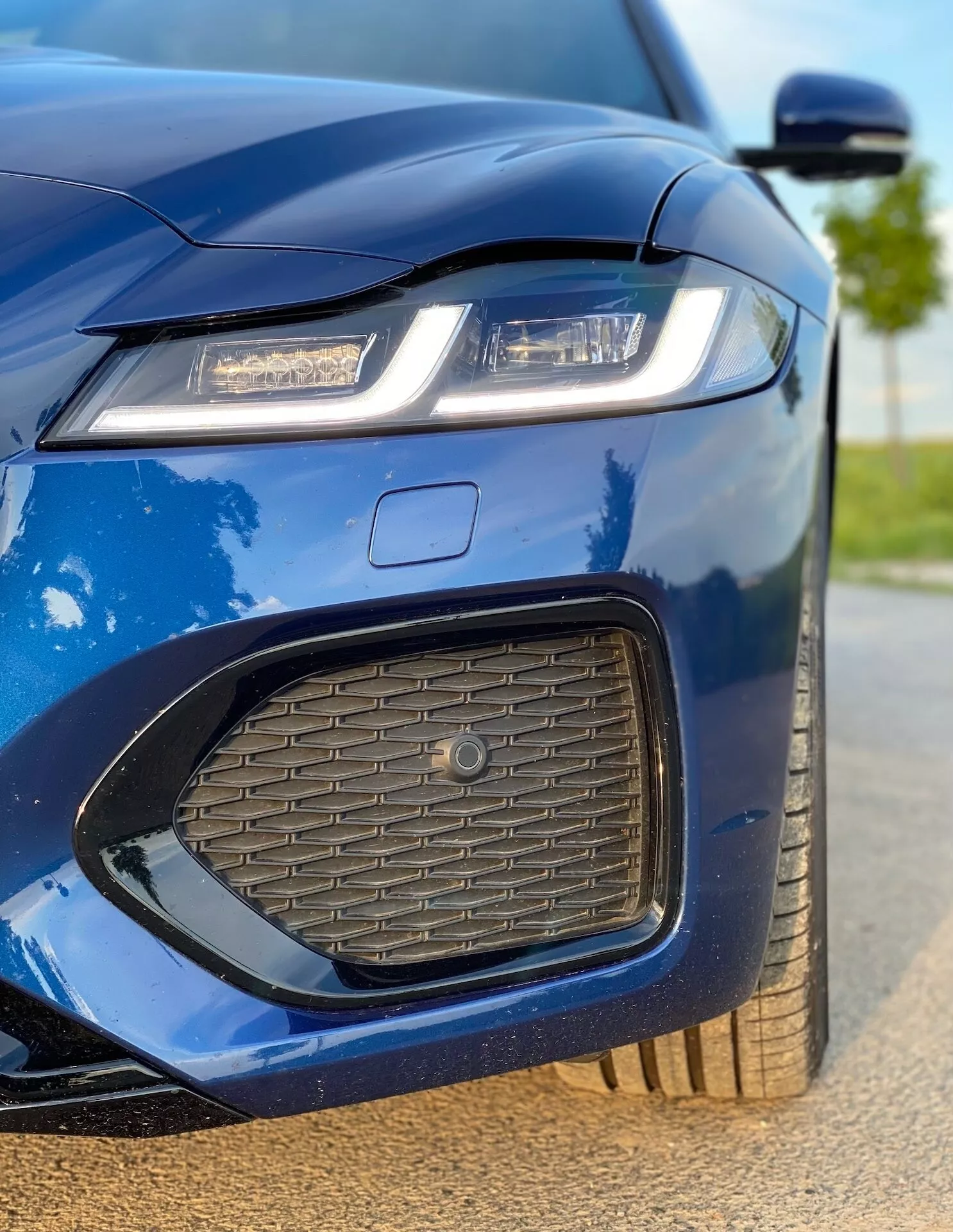 Zbliżenie przedniej lewej części Jaguara XF 2021 w metalicznym niebieskim kolorze. Zdjęcie uchwyca lewy reflektor z nowoczesną technologią LED lub laserową, część maski, przedni zderzak z wlotem powietrza o strukturze plastra miodu, część kratki z dużym emblematem oraz kawałek lewego lusterka bocznego. Obraz podkreśla ostry i agresywny design samochodu, sportową estetykę, wypolerowane wykończenie, aerodynamiczny kształt lusterka bocznego z wbudowanymi kierunkowskazami oraz częściowo widoczne aluminiowe koło, ukazując luksusowe i technologiczne cechy samochodu.