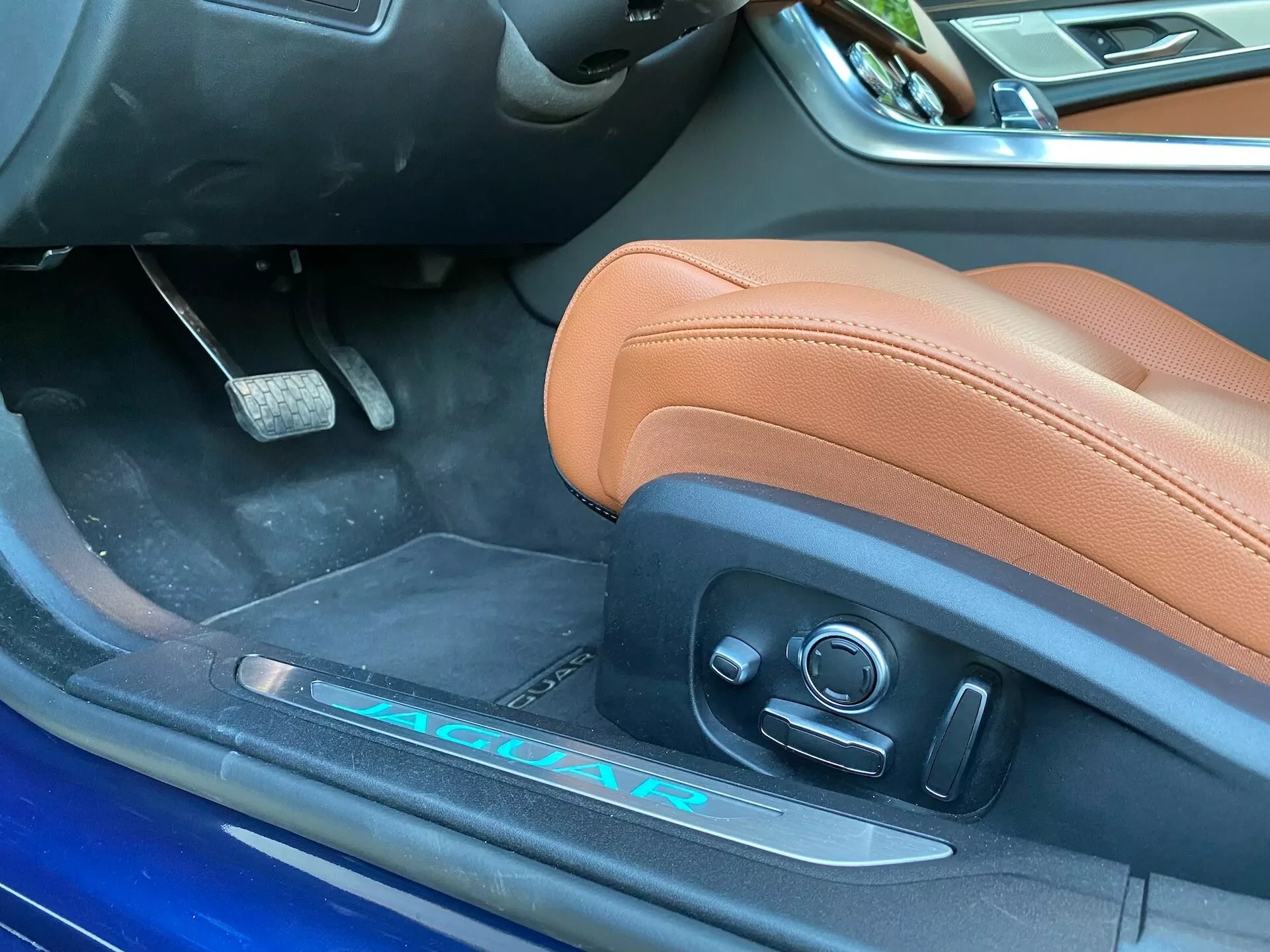Zbliżenie wnętrza po stronie kierowcy Jaguara XF 2021, ukazujące skórzane siedzenie kierowcy w kolorze jasnobrązowym z elektrycznymi regulatorami, częściowo widoczny aluminiowy próg drzwi z oznaczeniami 'JAGUAR' i 'R', oraz intensywnie niebieski brzeg drzwi. Zdjęcie ukazuje luksusowe i sportowe cechy pojazdu z lekko nachylonego kąta przy otwartych drzwiach.