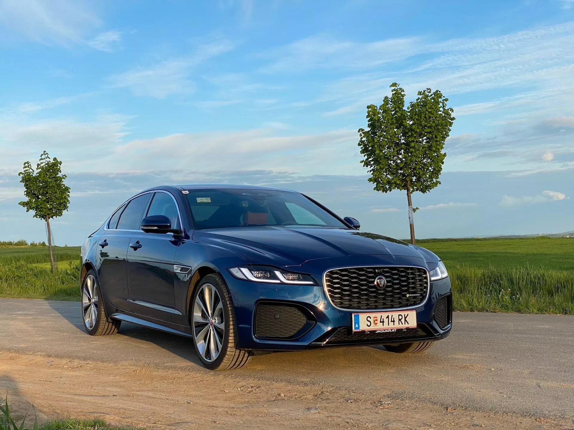 Na zdjęciu widać ciemnoniebieski luksusowy sedan Jaguar XF 2021 z niskiego przedniego kąta trzech czwartych. Pojazd stoi na żwirowym lub ziemistym wjeździe obok asfaltowej drogi, w tle trawa i czyste, błękitne niebo. Samochód ma reflektory LED i dużą, charakterystyczną przednią kratkę z centralnym znakiem sugerującym markę. Perspektywa trzech czwartych pokazuje maskę, przednią szybę, linię dachu oraz przedni boczny profil auta, prezentując felgi ze stopu metali z wieloramiennym wzorem. Maska z rzeźbionymi liniami, przyciemniane szyby dla prywatności i lusterka boczne z wbudowanymi kierunkowskazami sugerują dynamiczny i luksusowy design. Naturalne oświetlenie, połączone z widocznymi zaawansowanymi funkcjami technologicznymi, takimi jak systemy wspomagania kierowcy i opcje infotainment, podkreśla jego luksusowy status.