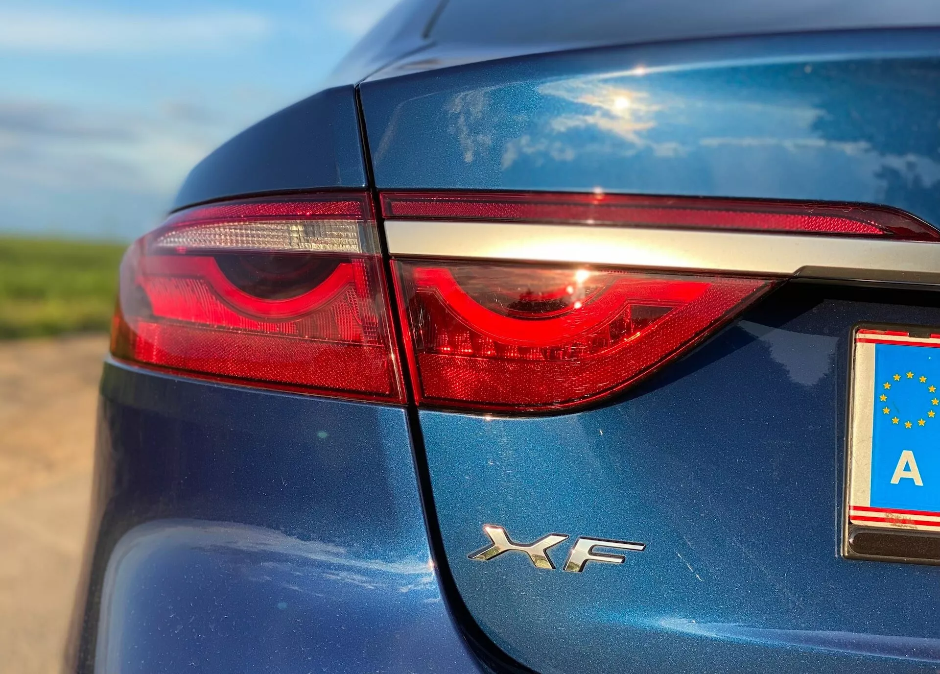 Zbliżenie przedstawiające tylnią, lewą część Jaguara XF 2021. Zdjęcie uwydatnia eleganckie, ciemnoniebieskie nadwozie, błyszczące wykończenie i lewe światło tylne z diodami LED. Tablica rejestracyjna UE ukazuje literę 'A', wskazującą na Austrię, z odbiciami świadczącymi o naturalnym świetle dziennym.