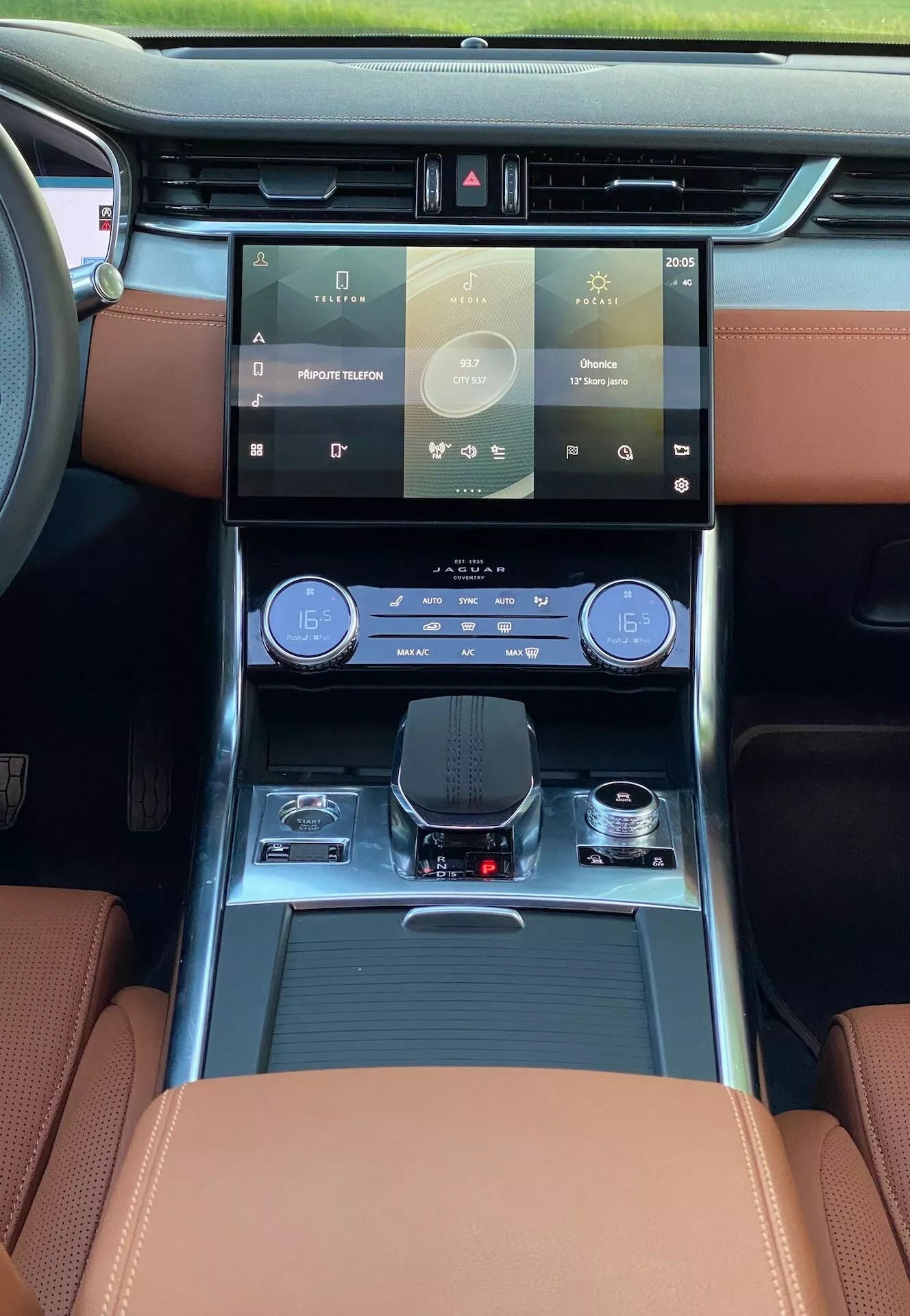 Wnętrze modelu Jaguar XF 2021, ukazujące perspektywę kierowcy z przedniego siedzenia, z naciskiem na deskę rozdzielczą i zaawansowane technologie. Kluczowe elementy obejmują duży, łatwy w obsłudze dotykowy system infotainment, elegancki panel dwustrefowej klimatyzacji, elektroniczny selektor biegów, luksusowe skórzane siedzenia z kontrastującymi przeszyciami i oświetlenie ambientowe. Widoczne są również praktyczne elementy, takie jak uchwyty na kubki i schowki. Naturalne światło zalewające kabinę sugeruje panoramiczny dach lub duże przednie okno, podkreślając ogólną luksusową atmosferę.