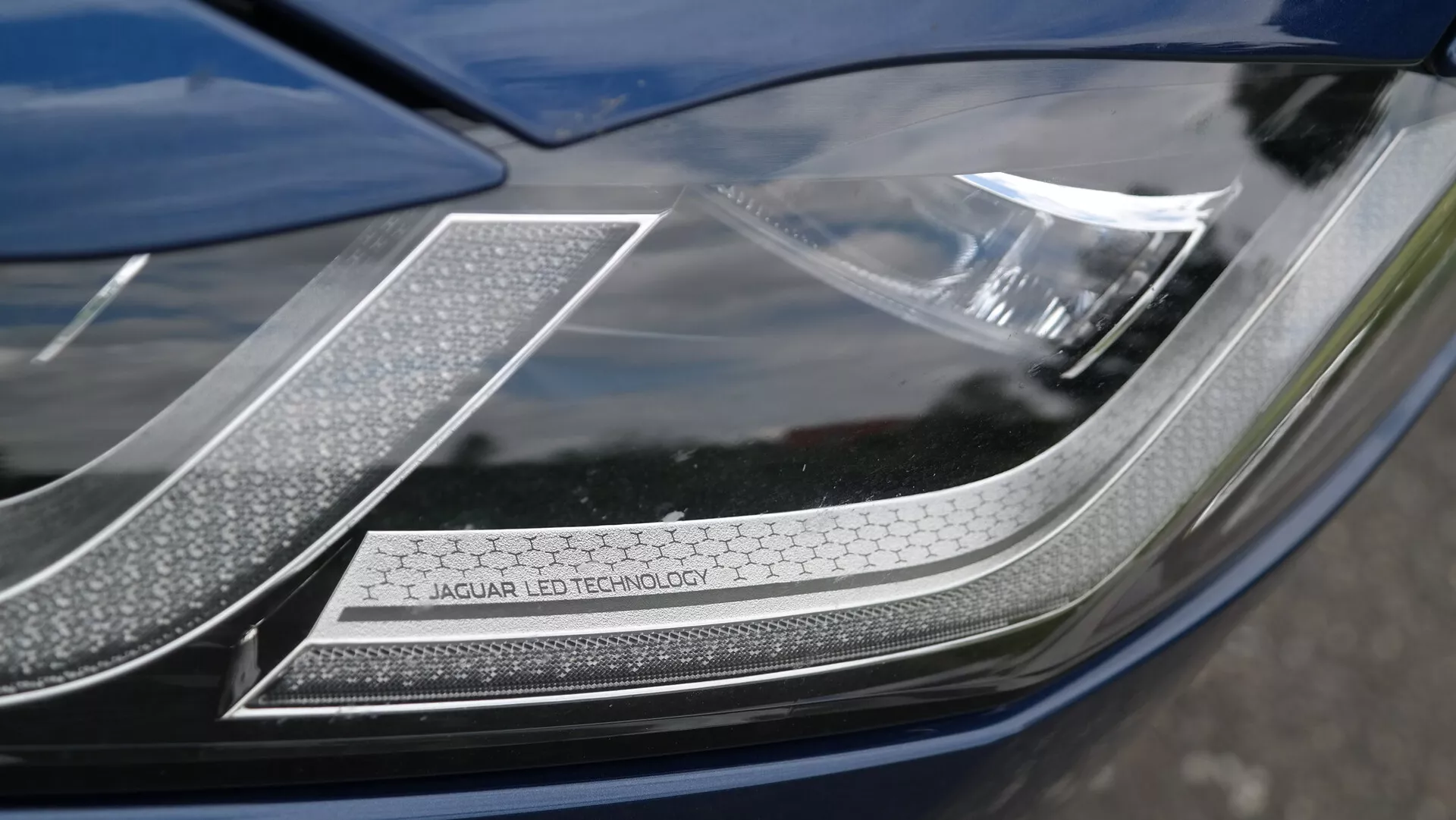 Zbliżenie reflektora samochodu Jaguar XF 2021, skierowane na prawy górny róg z lekko ukośnej perspektywy. Widoczne oznaczenie 'JAGUAR LED TECHNOLOGY' na chromowanej płytce, skomplikowany wzór diod LED i teksturowane światła do jazdy dziennej w kształcie plastra miodu. Reflektor posiada gładką, błyszczącą pokrywę z aerodynamicznym zakrzywieniem, podkreślającą zaawansowaną technologię LED i designerski wygląd premium Jaguara.