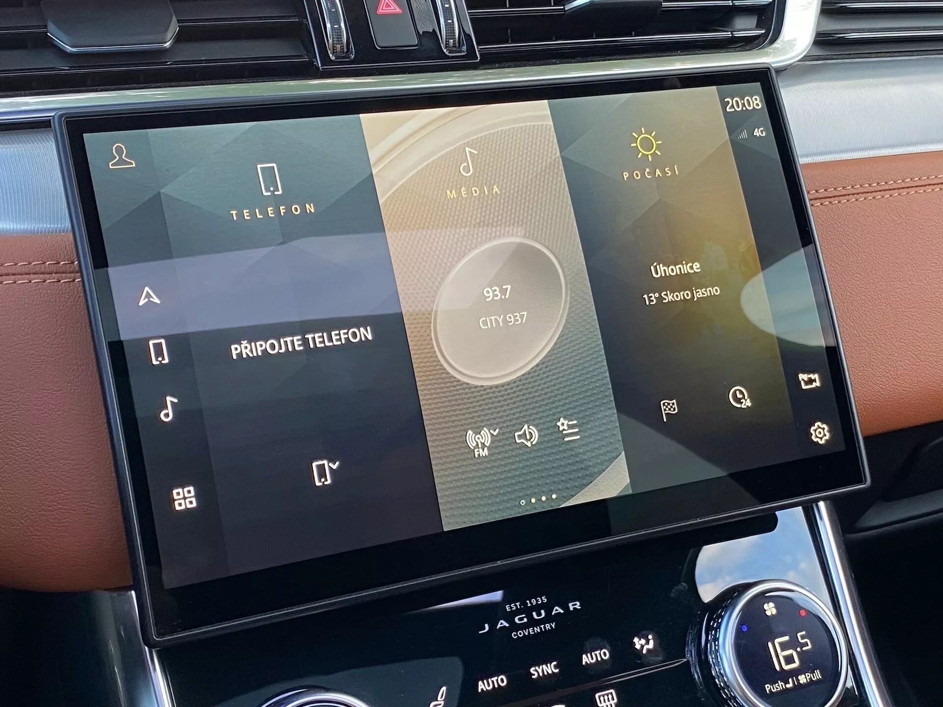 Dotykowy system infotainment samochodu Jaguar XF 2021 umieszczony w konsoli centralnej, widziany z perspektywy pasażera lub kierowcy. Skupienie na ekranie, wyświetlającym wiele ikon, takich jak łączność telefoniczna, odtwarzanie mediów i aplikacje specyficzne dla pojazdu, w tym Radio, Media i Podcasty, z oznakami integracji ze smartfonem. Nowoczesny interfejs dotykowy z eleganckim designem, pokazujący ustawienia systemu, takie jak czas (20:08) i AUTO dla klimatyzacji. Poniżej widoczny jest obrotowy pokrętło i dwustrefowe sterowanie klimatyzacyjne z ustawieniem temperatury na 16,5 stopnia Celsjusza.