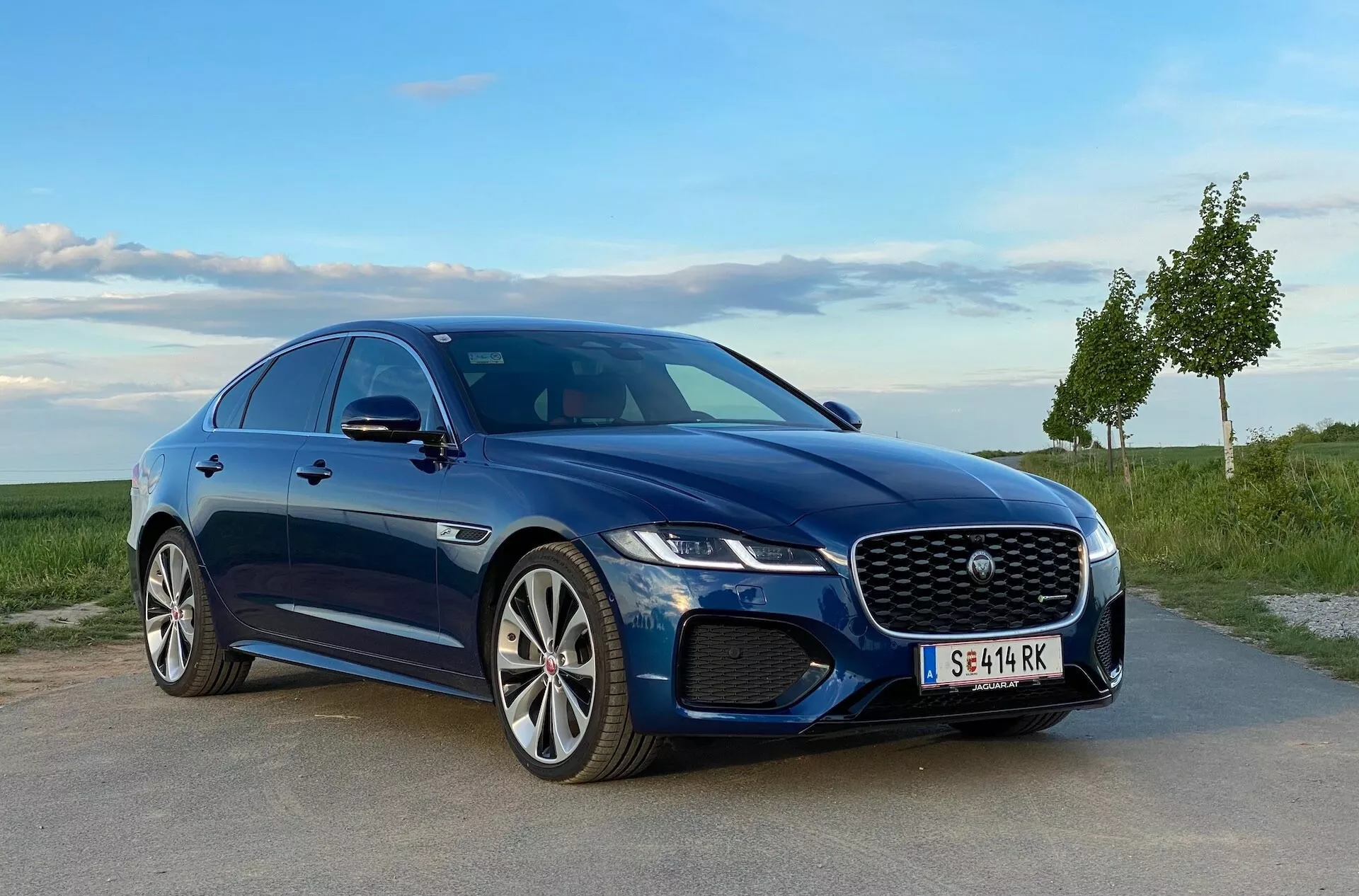 Niebieski sedan Jaguar XF 2021 zaparkowany na utwardzonej powierzchni, z trawiastym krajobrazem i czystym niebem w tle, pokazujący charakterystyczny grill, reflektory LED, stylowy przedni zderzak z wlotami powietrza i wieloramienne felgi aluminiowe. Sportowy profil samochodu jest podkreślony przez eleganckie linie i wycięte progi boczne, widziany z trzy czwarte przodu z niskiej perspektywy.
