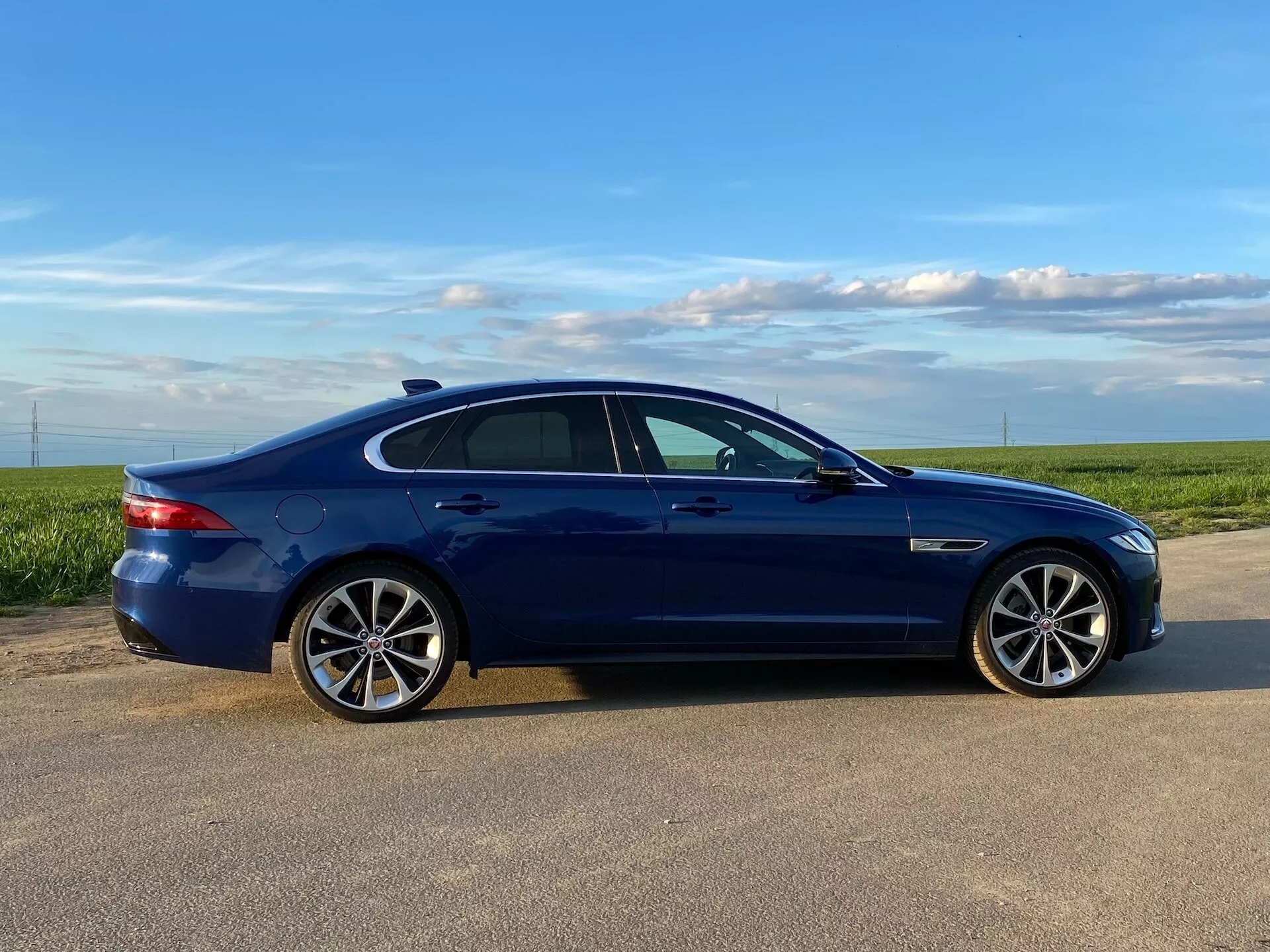 Widok z profilu bocznego niebieskiego sedana Jaguar XF 2021 uchwycony w dziennym świetle, pokazujący jego cechy projektowe, takie jak wieloramienne felgi aluminiowe, opony o niskim profilu, przyciemniane szyby, lusterka boczne w kolorze nadwozia z wbudowanymi kierunkowskazami, równo z karoserią kolorowe klamki drzwi i ozdobne listwy chromowane lub szczotkowane wzdłuż linii okien. Samochód jest ustawiony na utwardzonej powierzchni z otwartym polem i czystym niebem w tle, co sugeruje wiejskie lub półwiejskie otoczenie.
