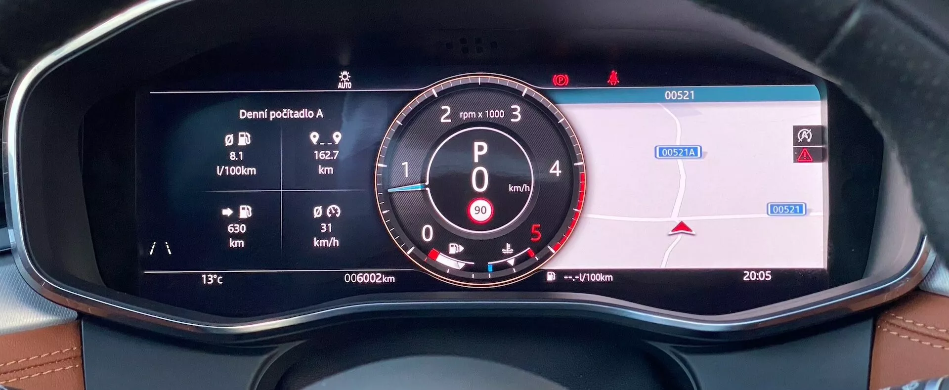 Zbliżenie widoku z fotela kierowcy cyfrowego zestawu wskaźników w Jaguarze XF 2021, wyświetlającego dane dotyczące zużycia paliwa, analogowy obrotomierz z RPM i cyfrowym prędkościomierzem, informacje nawigacyjne, odometr, temperaturę zewnętrzną i godzinę. Zestaw wskaźników jest obramowany deską rozdzielczą z przeszyciami z ekoskóry, podkreślając aspekty luksusowe i technologiczne wnętrza pojazdu.