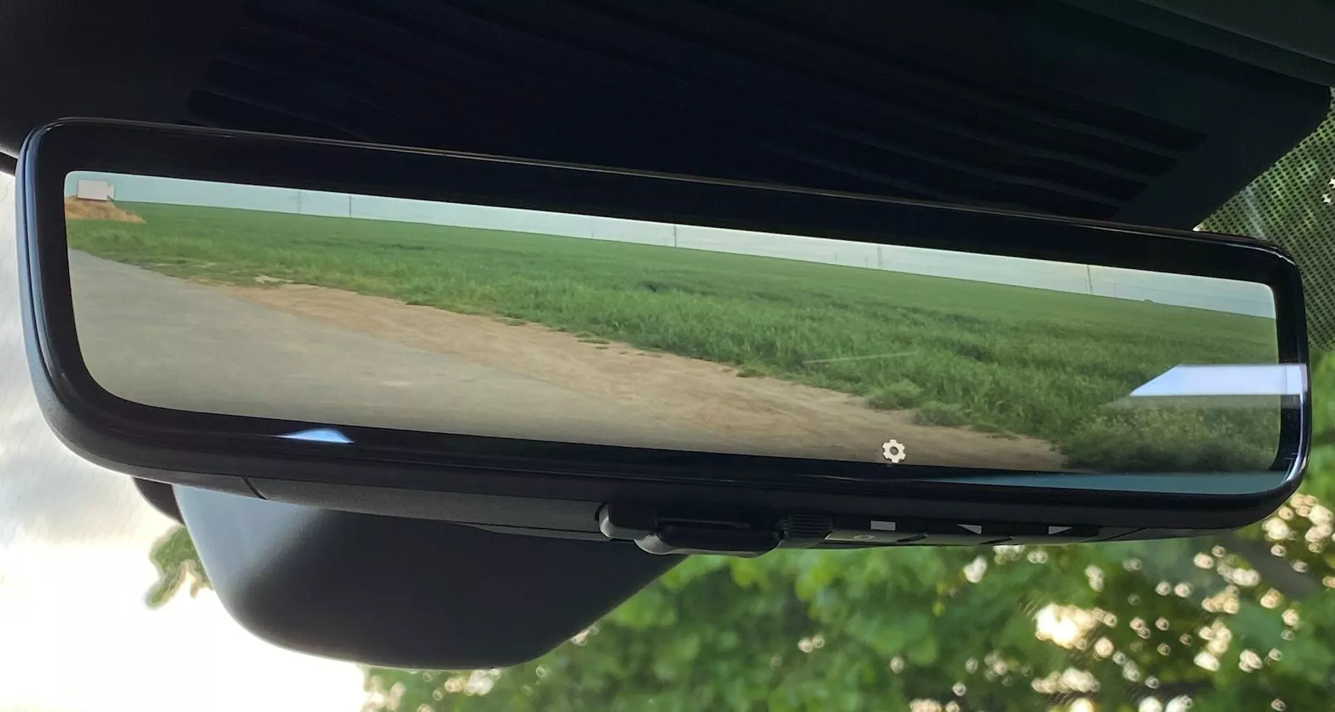 Lusterko wsteczne samochodu Jaguar XF z 2021 roku odzwierciedlające szerokokątny widok pola z roślinnością i czystym niebem, umieszczone centralnie na zdjęciu. Lusterko jest zamocowane na uchwycie szyby przedniej i otoczone czarną ramką, widoczne są sekcje podsufitki pojazdu i tylnego wnętrza.