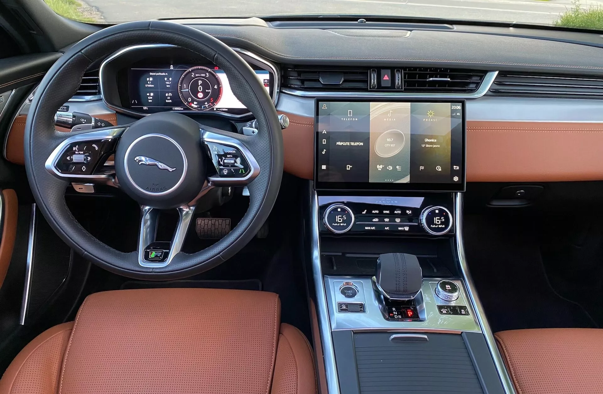 Wnętrze Jaguara XF 2021 widziane z miejsca kierowcy, prezentujące sportową kierownicę z płaskim spodem i wielofunkcyjnymi przyciskami, cyfrowy panel instrumentów, duży dotykowy system informacyjno-rozrywkowy, sterowanie klimatyzacją dwustrefową, przycisk bezkluczykowego uruchamiania, elektroniczny selektor biegów, nowoczesną konsolę środkową ze wstawkami drewnianymi i metalowymi oraz luksusową tapicerkę z karmelowej skóry.
