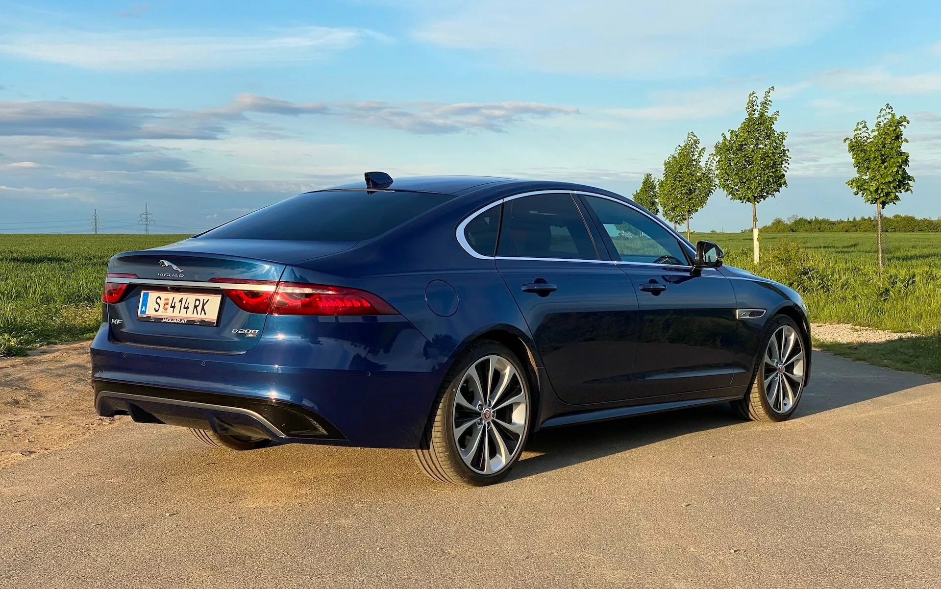 Widok z boku i tyłu nowoczesnego luksusowego sedana Jaguar XF 2021, sfotografowanego pod kątem trzy czwarte, koncentrując się na profilu bocznym i tylnym. Samochód stoi na żwirowym poboczu obok drogi z czystym niebem i zieloną roślinnością w tle. Cechy obejmują wydłużone światła tylne, które owijają się wokół boków, tylni spojler pokrywy bagażnika, wpuszczane klamki drzwi, okna z chromowanymi obramowaniami i felgi aluminiowe z wieloramiennym wzorem.