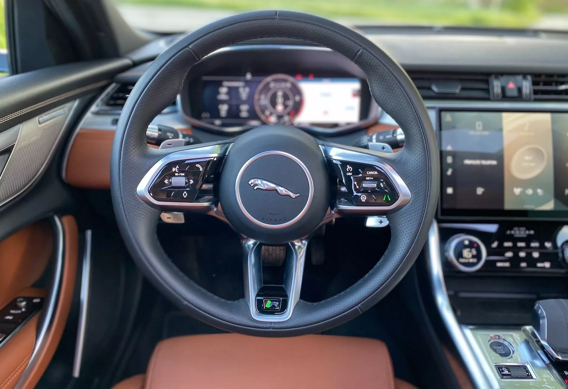 Widok wnętrza Jaguar XF 2021 z perspektywy siedzenia kierowcy, ukazujący czarną, skórzaną, wielofunkcyjną kierownicę ze srebrnym emblematem i zaawansowanymi przyciskami sterującymi. W pełni cyfrowy zestaw wskaźników za kierownicą posiada wyświetlacz o wysokiej rozdzielczości. Deska rozdzielcza z luksusowymi drewnianymi wstawkami rozciąga się na boki. Na środku widać górną krawędź dotykowego systemu informacyjno-rozrywkowego i panelu kontroli klimatu, co wskazuje na zaawansowane technologie i luksusowe cechy pojazdu.