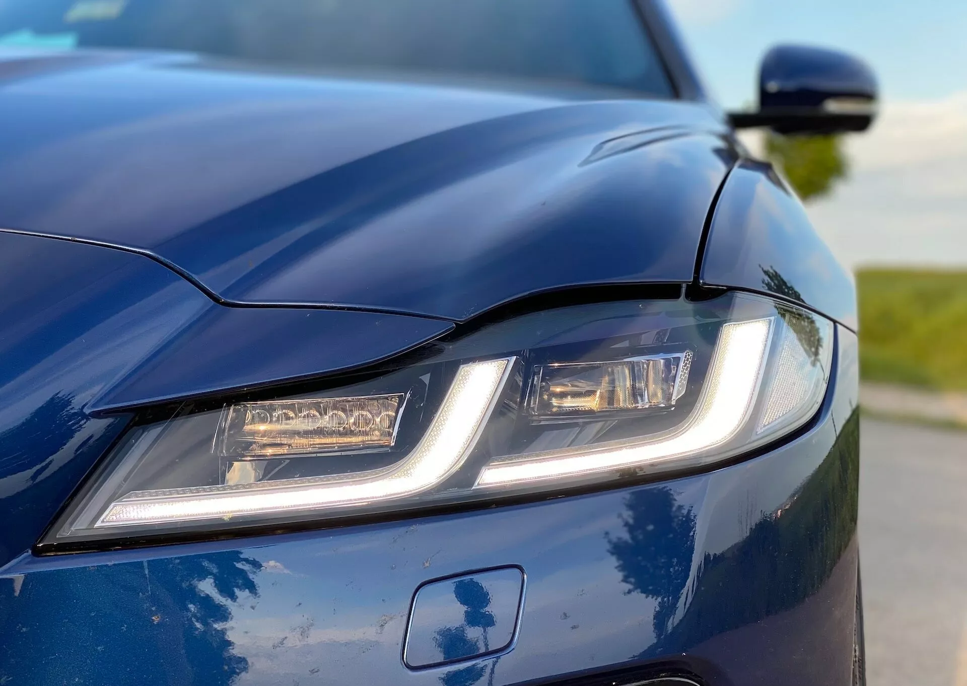 Zbliżenie przedniej, lewej sekcji Jaguara XF 2021 z niskiego, lekko przesuniętego kąta, podkreślające nowoczesny lewy reflektor z charakterystycznymi dziennymi światłami LED. Głęboki niebieski lakier i rzeźbiona maska ​​z aerodynamicznymi liniami są wyraźnie widoczne. Częściowo widoczne jest również lusterko poboczne, co dodaje eleganckiego wyglądu. Płytka głębia ostrości podkreśla design samochodu i zaawansowany system oświetleniowy.