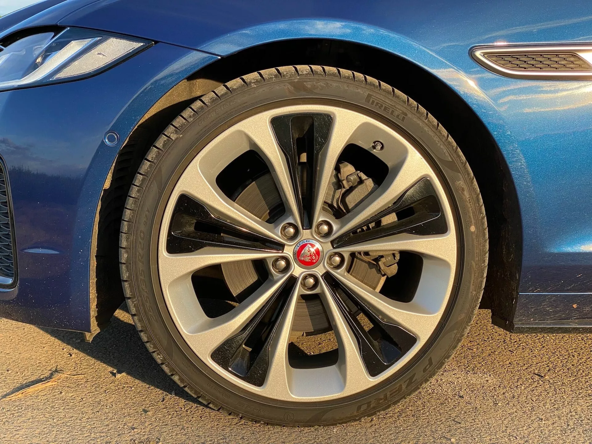 Zbliżenie przedniego koła pasażera Jaguara XF 2021 z niskiego kąta. Wieloramienna felga aluminiowa ma dynamiczny design z dziesięcioma parami cienkich i grubych ramion. Środek felgi ma czerwone logo marki. Opona drogowa ma widoczny bieżnik, a za ramionami felgi widać zacisk hamulcowy i wentylowany tarcz hamulcowy. Widać część niebieskiego nadwozia z błyszczącym wykończeniem oraz zakrzywiony błotnik. Obraz podkreśla luksusowe i stylowe cechy samochodu, odpowiedni do dyskusji na temat części samochodowych, detali projektowych i identyfikacji marki.