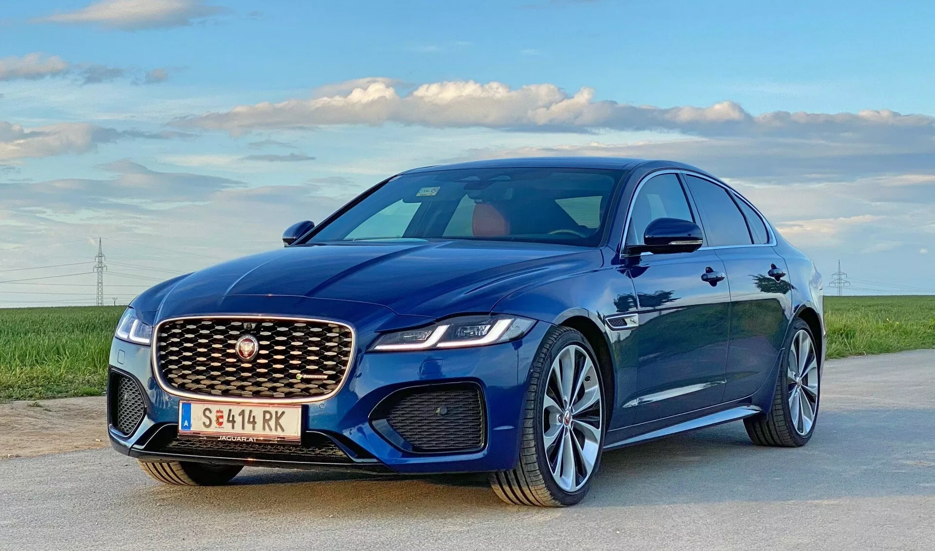 Obraz przedstawia nowoczesny luksusowy sedan, Jaguar XF 2021, zaparkowany na otwartej drodze pod czystym niebem z polami i odległymi liniami energetycznymi na horyzoncie. Ujęcie z niskiego kąta i z trójwymiarowym widokiem z przodu podkreśla stronę kierowcy. Wyróżnia się wyraźny grill z siatkowym wzorem i centralnym emblematu, eleganckie reflektory LED, rzeźbione linie maski, aerodynamiczne lusterka boczne oraz wieloszprychowe felgi aluminiowe z metalicznym lakierem. Można założyć obecność zaawansowanych reflektorów LED, aktywnej aerodynamiki, felg aluminiowych z oponami o wysokich osiągach oraz potencjalnie zaawansowanych systemów wspomagania kierowcy.