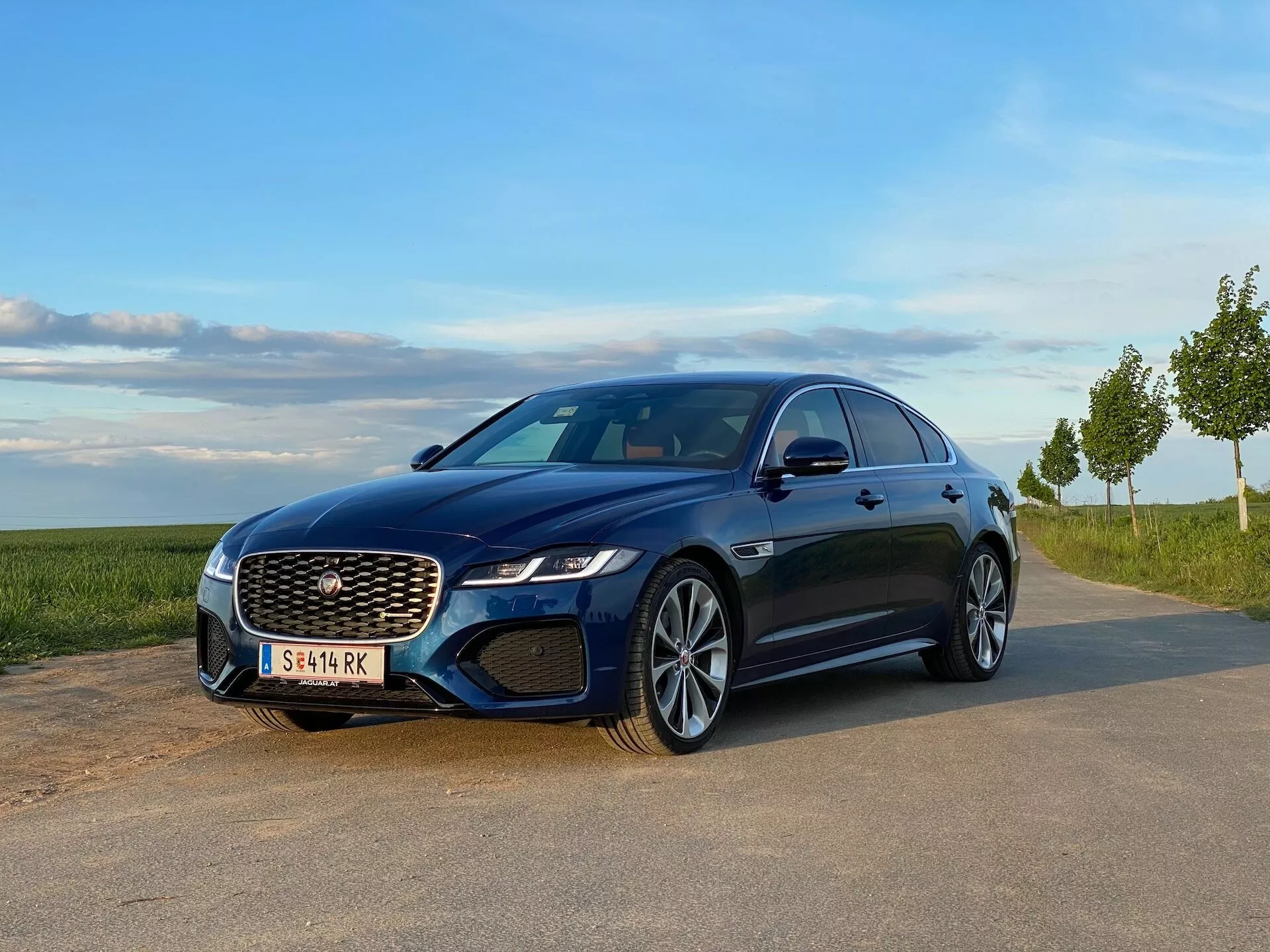 Niebieski sedan Jaguar XF 2021 zaparkowany na utwardzonym terenie z naturalnym krajobrazem w tle, uchwycony z widoku z przodu pod kątem trzech czwartych. Widoczne cechy obejmują wyróżniającą się kratkę z wzorem siatki, kątowe reflektory LED, zintegrowane światła przeciwmgielne, wieloszprychowe felgi aluminiowe, elegancką linię dachu i boczne panele z wyraźnymi liniami charakteru, pokazujące nowoczesne i sportowe elementy wzornictwa.