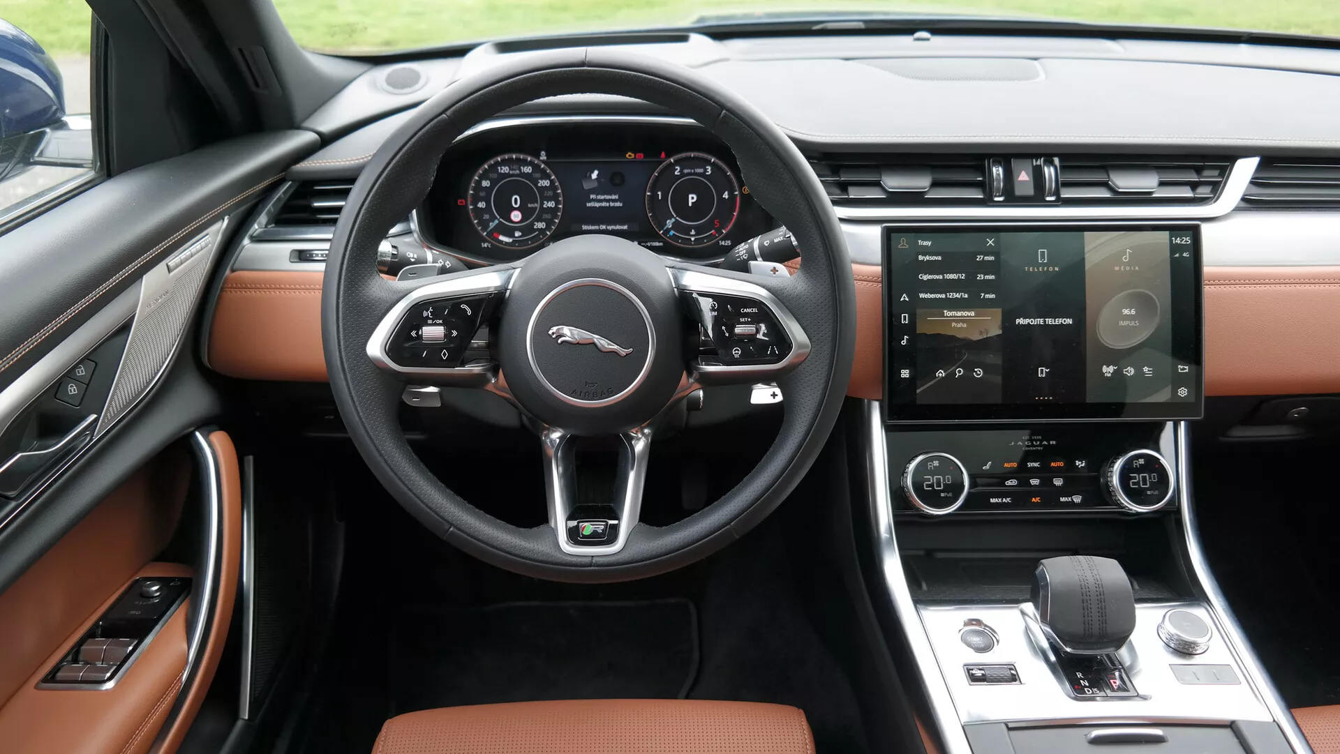 Wnętrze Jaguara XF 2021 skierowane na przednią część kabiny z centralnie umieszczoną, trójramienną kierownicą z funkcjami wielozadaniowymi. Cyfrowy zestaw wskaźników wyświetla informacje o pojeździe. Konsola środkowa zawiera duży system infotainment z ekranem dotykowym oraz system klimatyzacji dwustrefowej. Obok znajduje się elektroniczny selektor obrotowy oraz eleganckie wnętrze premium w dwóch odcieniach – czarnym i brązowo/beżowym. Panel drzwiowy i częściowo widoczne fotele sugerują tapicerkę skórzaną lub skórzanopodobną oraz elektryczne szyby.