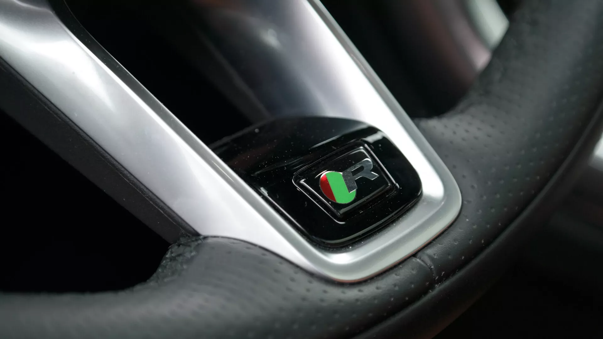 Zbliżenie widoku z perspektywy kierowcy na kierownicę Jaguara XF 2021, z cyfrowym wyświetlaczem w centrum pokazującym flagę wyścigową, sugerującą aktywowany tryb sportowy. Obraz podkreśla luksusowy skórzany materiał z widocznymi szwami i teksturą z włókna węglowego, uzupełnione chromowanym akcentem wokół ekranu, odbijającym jasne oświetlenie.