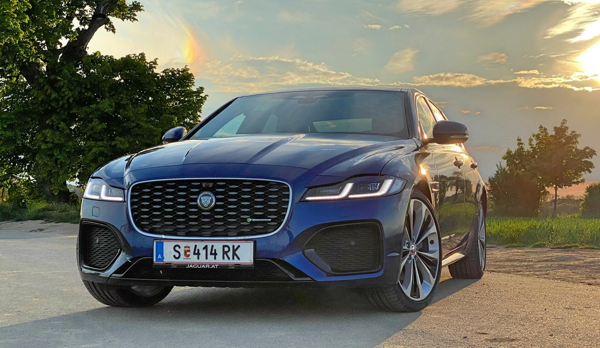 Obraz przedstawia Jaguara XF 2021 z niskiego kąta trzy czwarte przód, podkreślając jego aerodynamiczny design, dużą kratkę z emblematem marki, ostre reflektory LED, wyraźny zderzak z dużymi wlotami powietrza, rzeźbione linie maski i elegancki profil boczny. Samochód ma duże wieloramienne felgi aluminiowe, lusterka boczne z wbudowanymi kierunkowskazami, charakterystyczne linie nadwozia i lekko przyciemniane szyby. Ustawiony na drodze lub parkingu z malowniczym tłem drzew i niebem z widocznym zachodem słońca, pojazd emanuje siłą i ruchem do przodu.