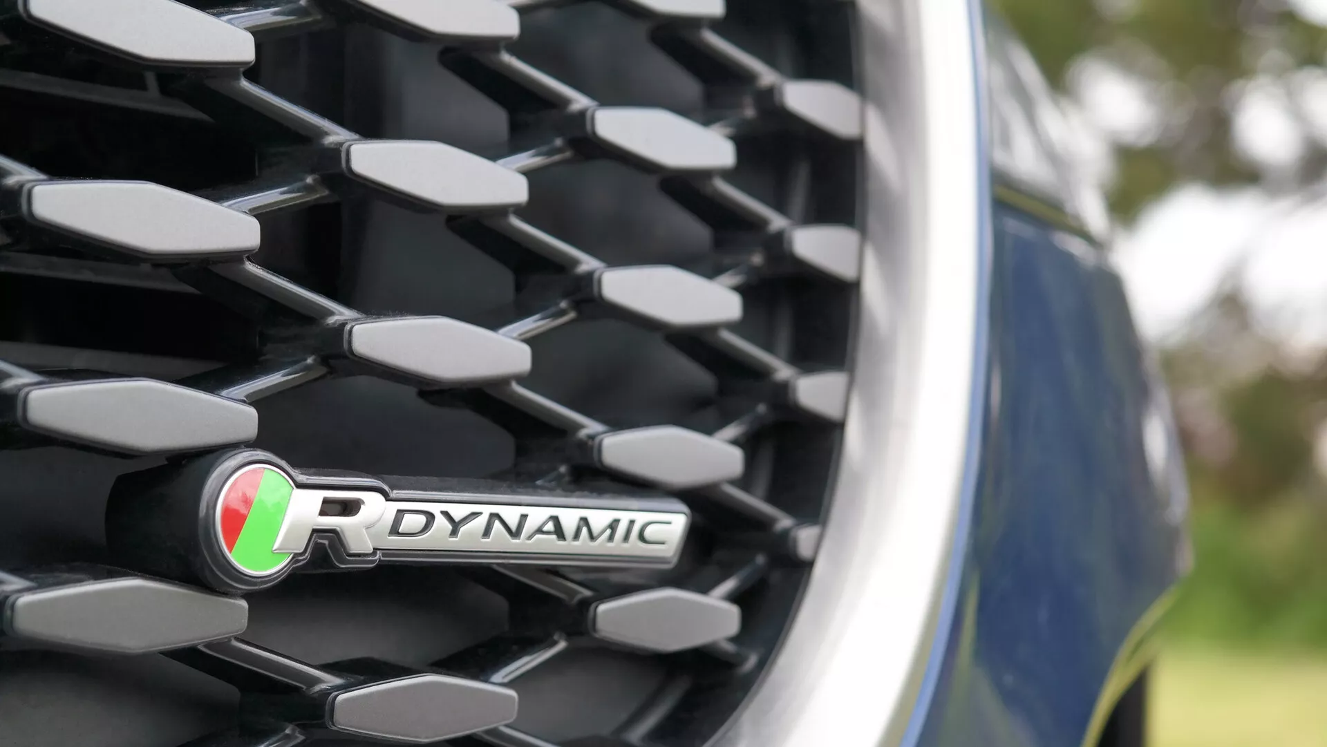 Zbliżenie na przedni grill Jaguara XF R-Dynamic z 2021 roku o wzorze diamentowym/plaster miodu, chromowane wykończenie i widoczny znaczek z zielonymi i czerwonymi paskami. Częściowo widoczny błyszczący niebieski lakier. Plener z rozmazaną zielenią w tle.