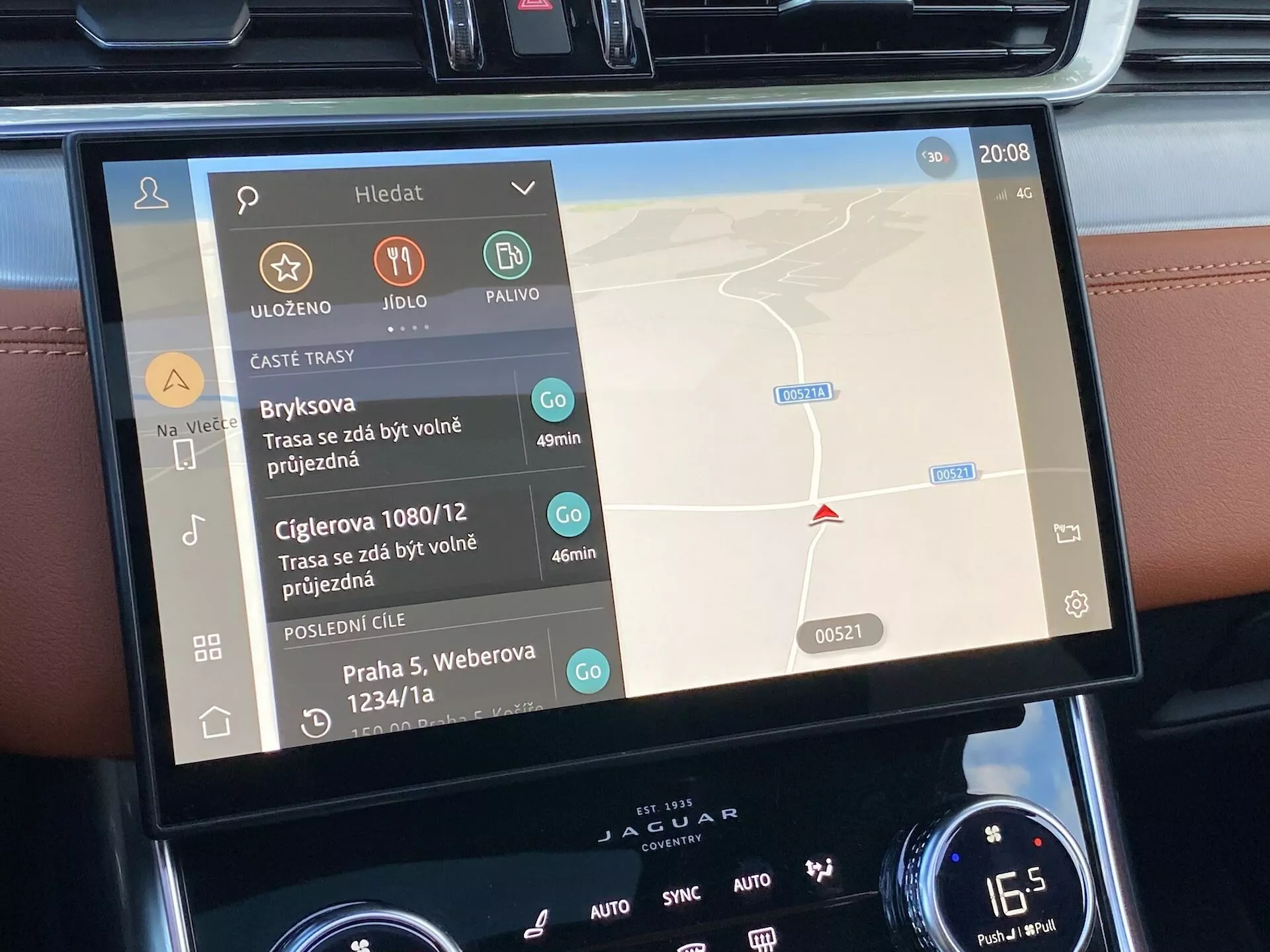 Widok wnętrza wyświetlacza systemu informacyjno-rozrywkowego w Jaguarze XF 2021, pokazujący dwujęzyczny interfejs dotykowy z mapą nawigacji po prawej stronie i kontrolkami multimediów po lewej, otoczony przyciskami i pokrętłami klimatyzacji, wskazując na luksusowe funkcje technologiczne.