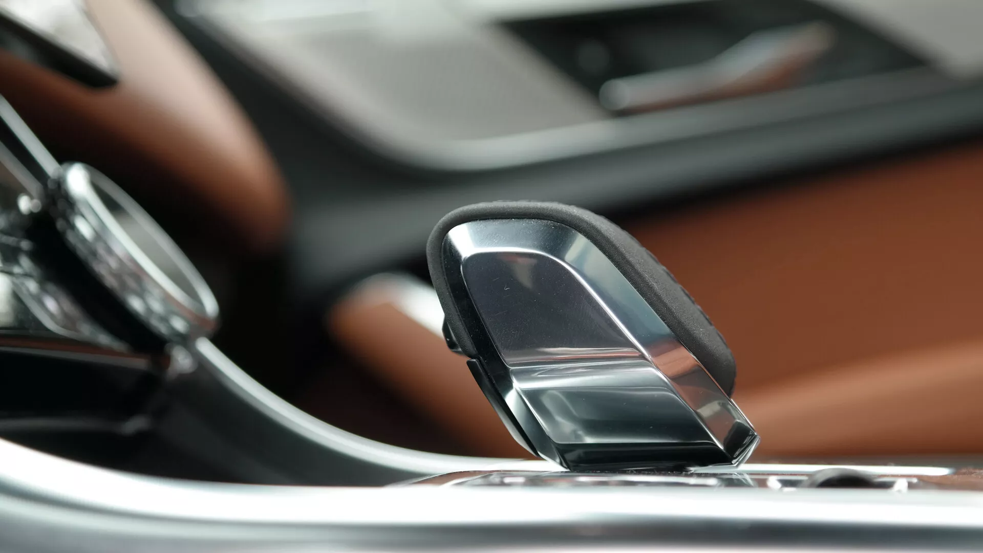 Zbliżenie wnętrza Jaguara XF 2021, skupiające się na eleganckim joysticku lub pokrętle zmiany biegów. Perspektywa jest nieco wyższa i skierowana w dół, podkreślając dźwignię zmiany biegów na pierwszym planie. Po lewej stronie widoczna jest część okrągłego pokrętła sterującego związanego z systemem infotainment lub audio. Deska rozdzielcza, wykończona luksusowym drewnem, jest widoczna za dźwignią zmiany biegów, z ekranem dotykowym zintegrowanym z konsolą centralną z metalicznymi akcentami w tle. Rozmyte tło podkreśla dźwignię zmiany biegów, sugerując małą głębię ostrości. To sugeruje automatyczną skrzynię biegów i nowoczesny system infotainment, wskazując na wysokiej klasy luksusowy pojazd.