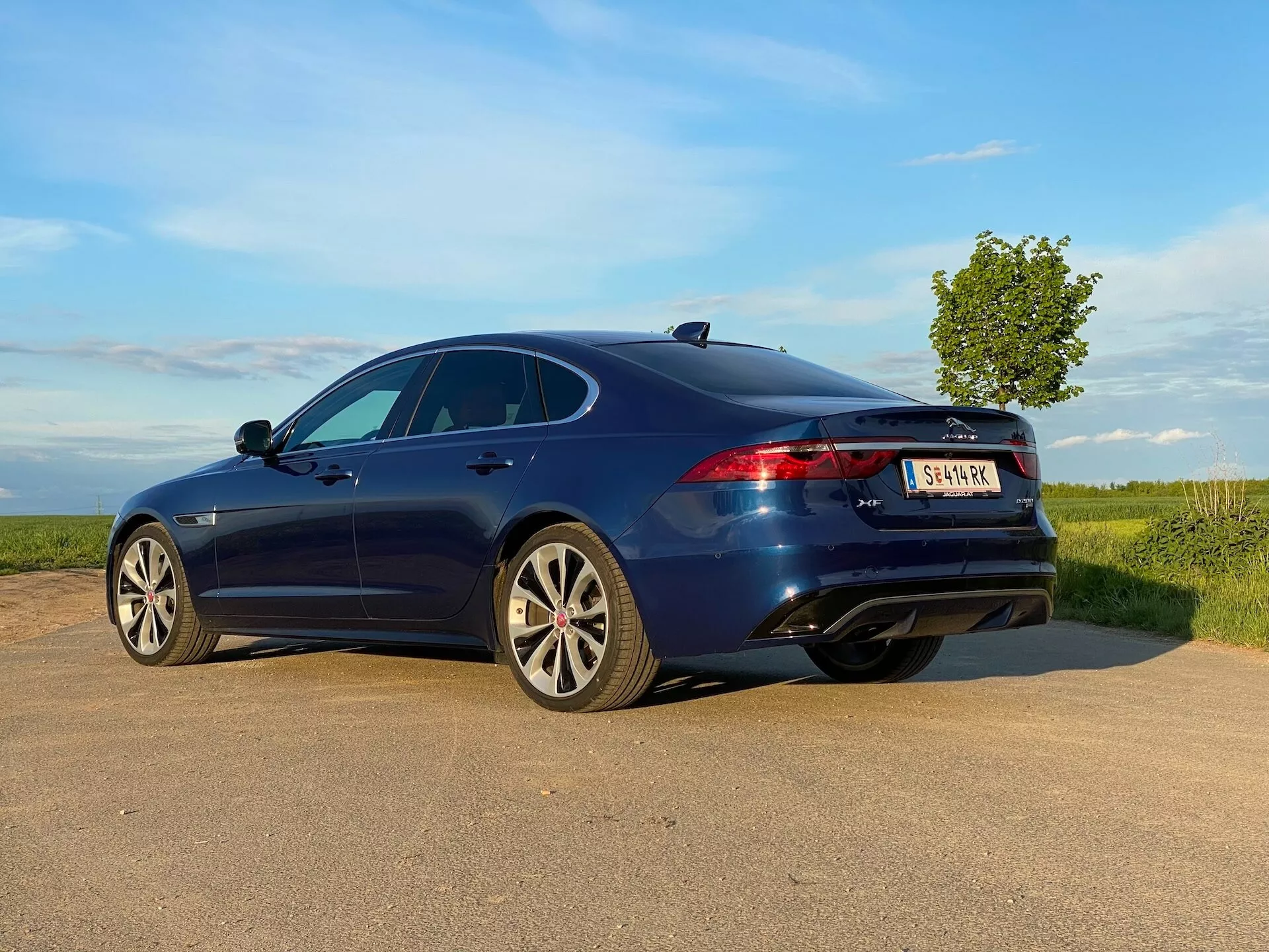 Nowoczesny czterodrzwiowy sedan w głębokim niebieskim kolorze widziany z tyłu i z boku na wiejskiej drodze. Pojazd ma elegancki, aerodynamiczny profil z fastback designem i aluminiowymi felgami o wieloszprychowym wzorze. Obraz pokazuje również szyberdach, nowoczesne tylne światła LED, trzecie światło stopu i podwójne rury wydechowe. Naturalne, częściowo pochmurne niebo w tle sugeruje otwarte, wiejskie otoczenie, co dodaje dynamiczności zdjęciu wykonanym pod niskim kątem.