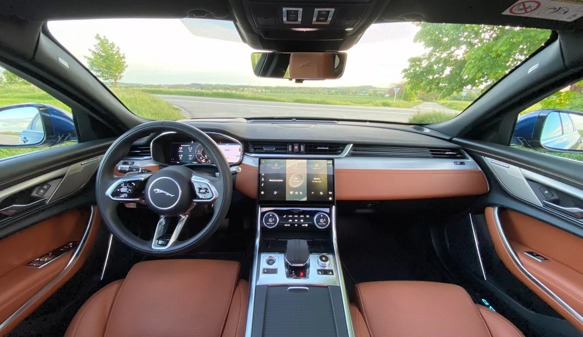 Widok wnętrza Jaguara XF 2021 z perspektywy tylnego siedzenia, prezentujący luksusowe brązowe skórzane siedzenia, wielofunkcyjną kierownicę z przyciskami, cyfrowy zestaw wskaźników, podwójne ekrany dotykowe do systemu infotainment i klimatyzacji, elektronikę do zmiany biegów oraz wyrafinowany design deski rozdzielczej z zintegrowanymi nawiewami.